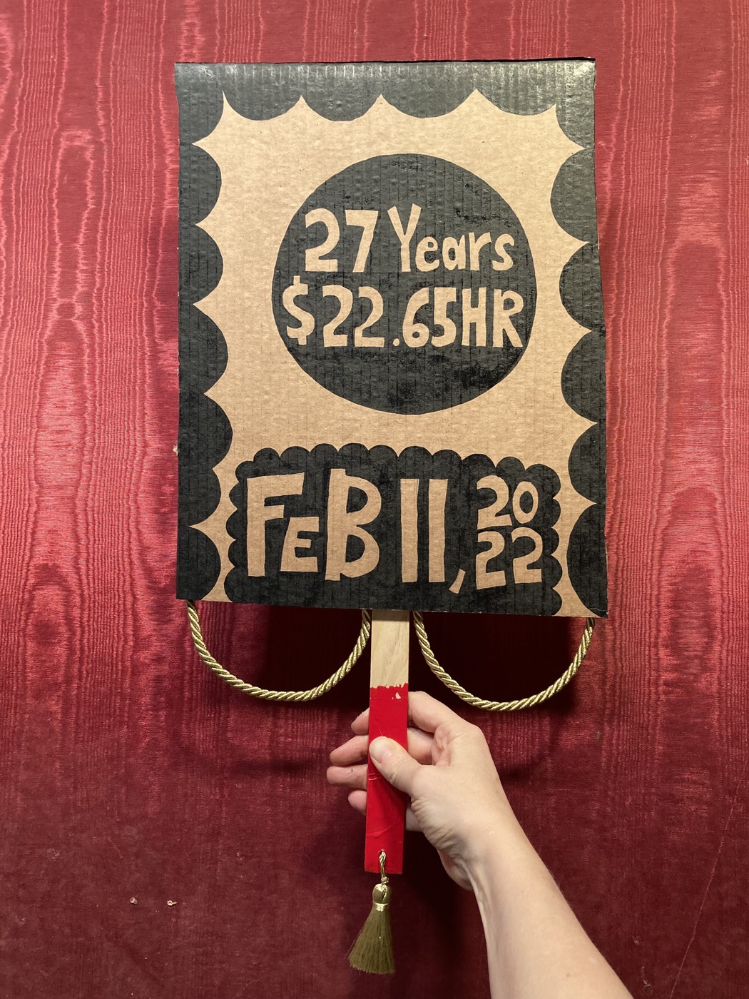 A cardboard picket sign is held by a light-skinned arm against a maroon, textured wall. It reads "27 Years, $22.65/hr" in a black circle and below that "Feb 11, 2022." It is decorated in a scalloped black-ink border. The sign's handle is painted red and has a gold tassel hanging from its bottom.