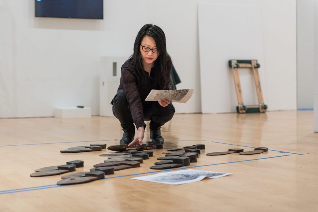 In a contemporary museum gallery, a light-skinned woman of Asian descent crouches as she arranges unidentified, flat, black sculptures on the floor, all within a blue, taped-off area. She holds a piece of paper that she seems to be referencing and another paper is laid out on the floor a ways in front of her. She wears all black and black-framed glasses.