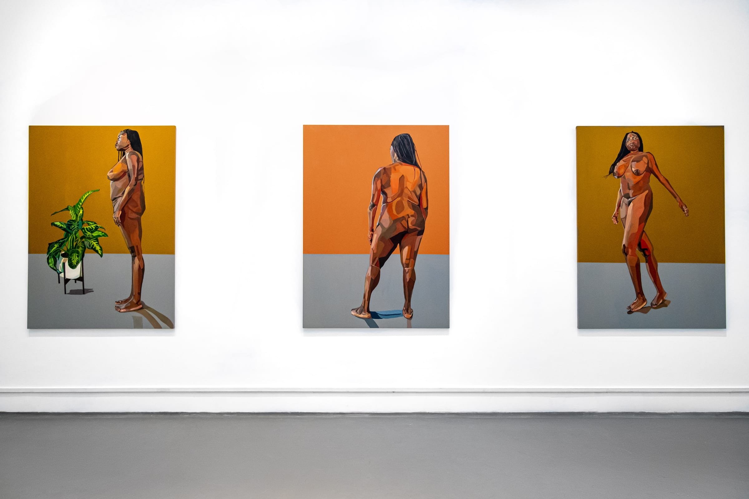 Three vertical, earth-toned, figurative paintings hang on a white gallery wall. Each depicts a nude woman with medium-dark skin standing in different postures: to the side, with her back to the viewer, and in motion turning around. The first painting also includes a lush green plant in a white pot.