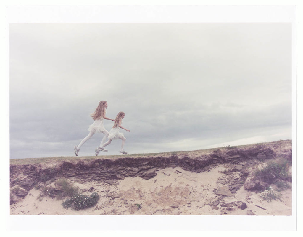 A photograph shows two young, twin girls running on a dirt road in a sparse, cloudy landscape. They have long, blonde hair and wear identical white leggings and a sleeveless white dress with a ruffled skirt. They are captured mid-stride.