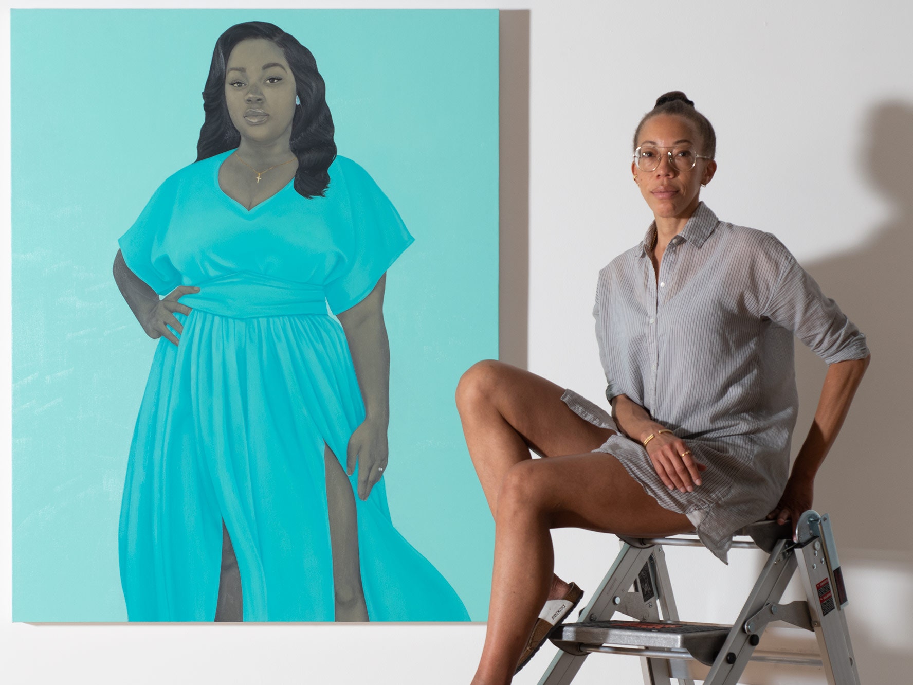 A medium-dark skinned woman sits on an industrial folding ladder next to a large, ethereal, painted portrait of a young woman whose skin is rendered in greyscale. The subject wears a flowing, light blue dress and stands confidently, one hand on her hip, with a kind and knowing gaze fixed on the viewer. The painting's background is also light blue.