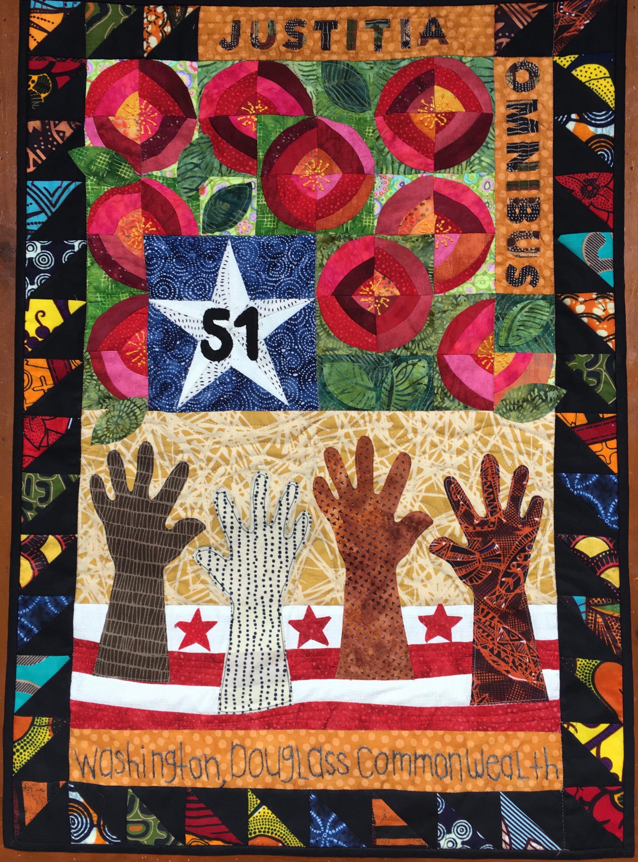 A vertical patchwork quilt features many colorful squares and four brown, white, and patterned hands reaching up from the bottom towards a blue-patterned square that holds a white star and the number "51" centered within it, in black.