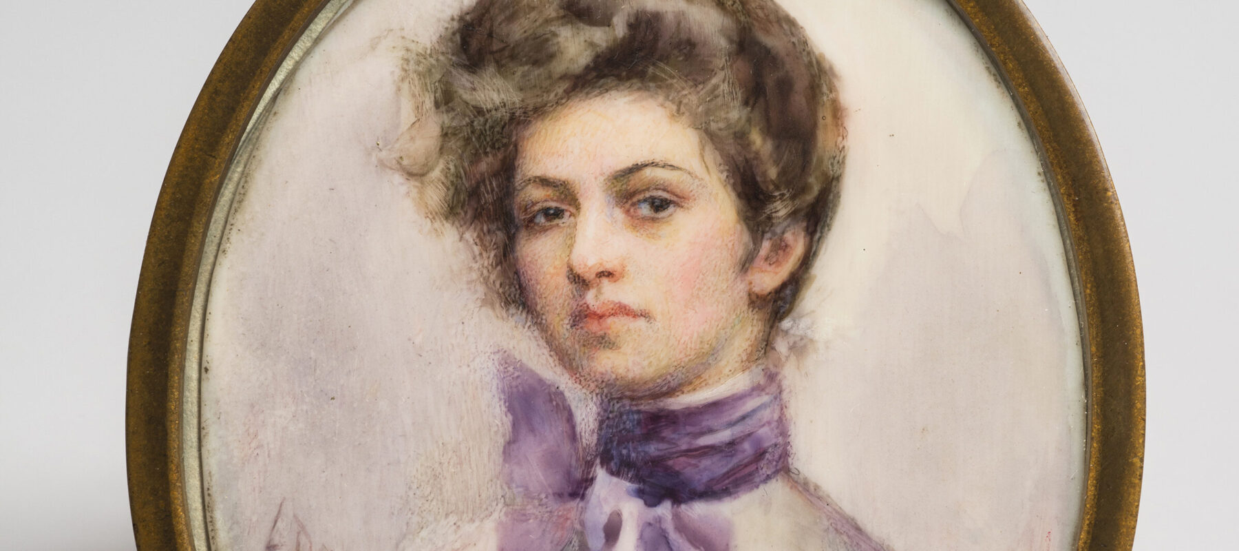 Miniature half-portrait of a light-skinned woman, set in an oval-shaped brass frame, gazing confidently at the viewer.  The woman is wearing a Victorian-style white blouse with a purple scarf tied in bow around her neck, her brown hair loosely piled atop her head.
