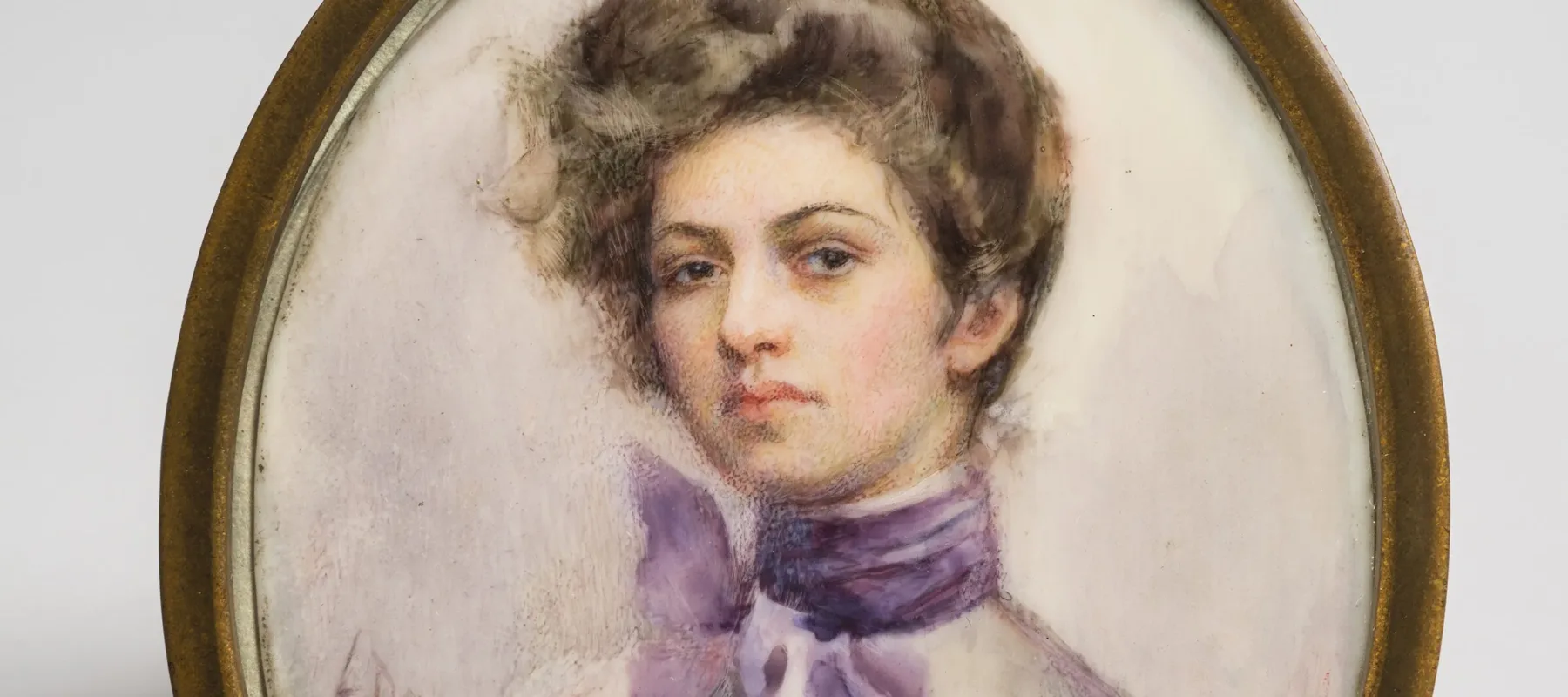 Miniature half-portrait of a light-skinned woman, set in an oval-shaped brass frame, gazing confidently at the viewer.  The woman is wearing a Victorian-style white blouse with a purple scarf tied in bow around her neck, her brown hair loosely piled atop her head.
