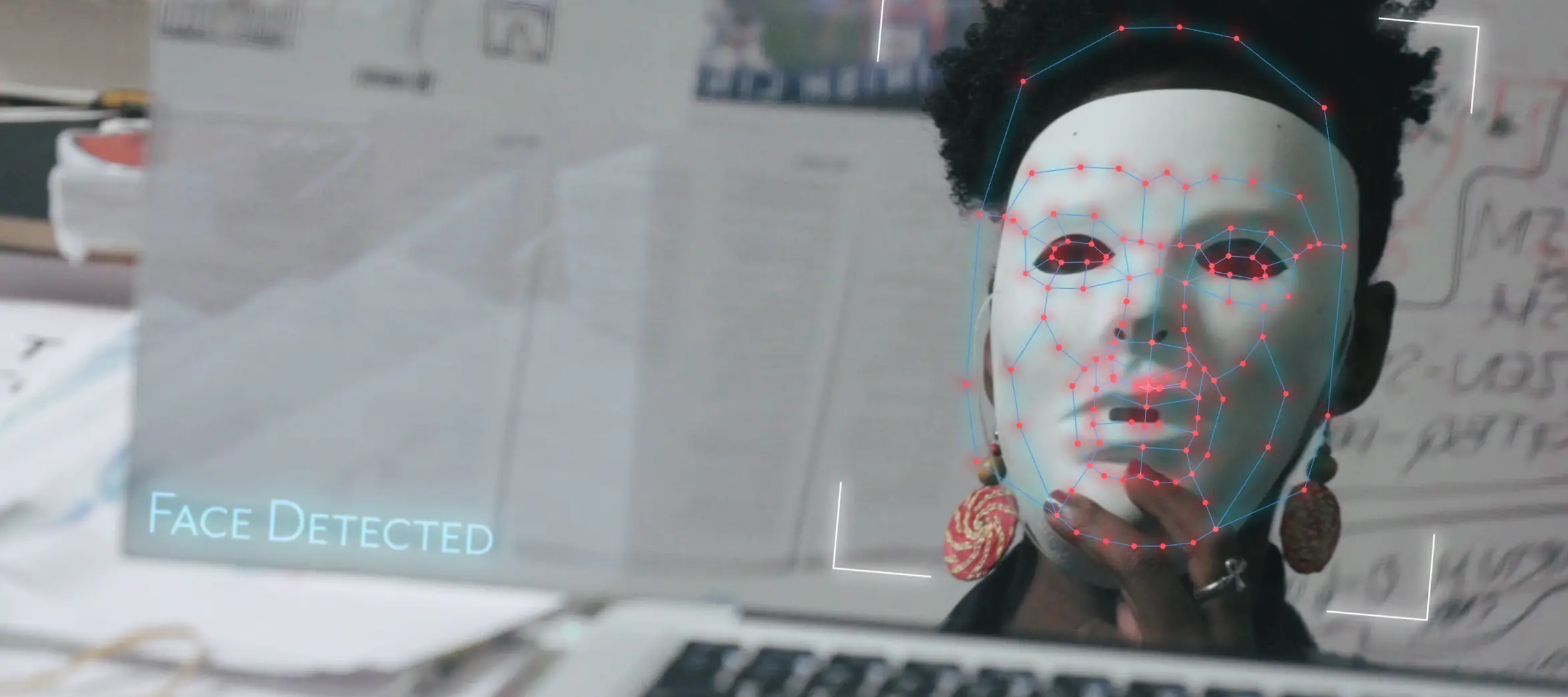 A computer screen shows a dark-skinned adult woman with short, dark, bushy hair holds up a smooth, white mask to obscure her face. Layered over the mask are a constellation of red dots connected by blue lines, and in the corner of the screen are the words ‘Face Detected.’