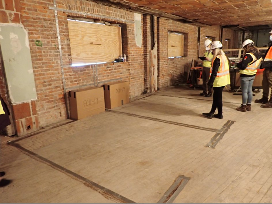 Four people in hard hats and safety vests look at the outline of an office marked up on the bare floor on the gutted office area.