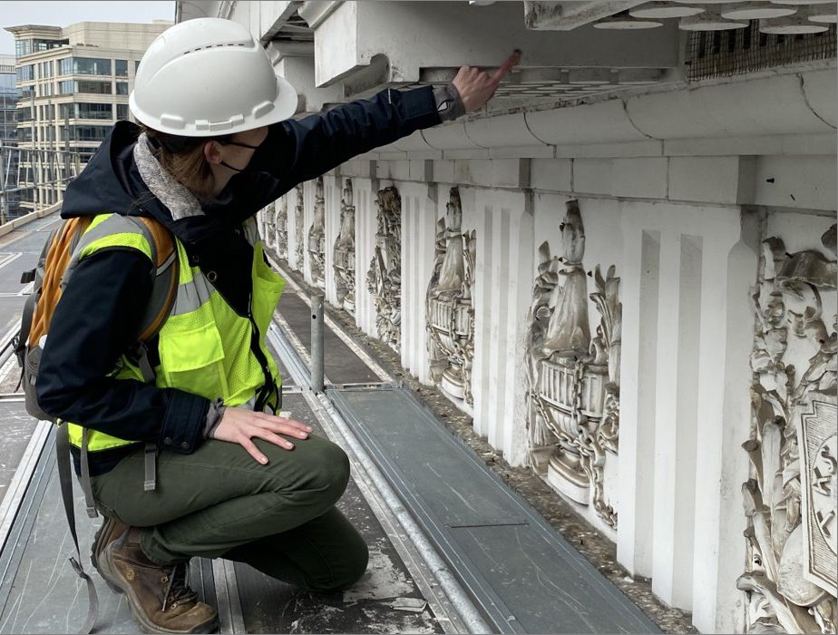 Individual with light skin wearing a white hard had and florescent green vest inpsects the decorative cornice of the building.