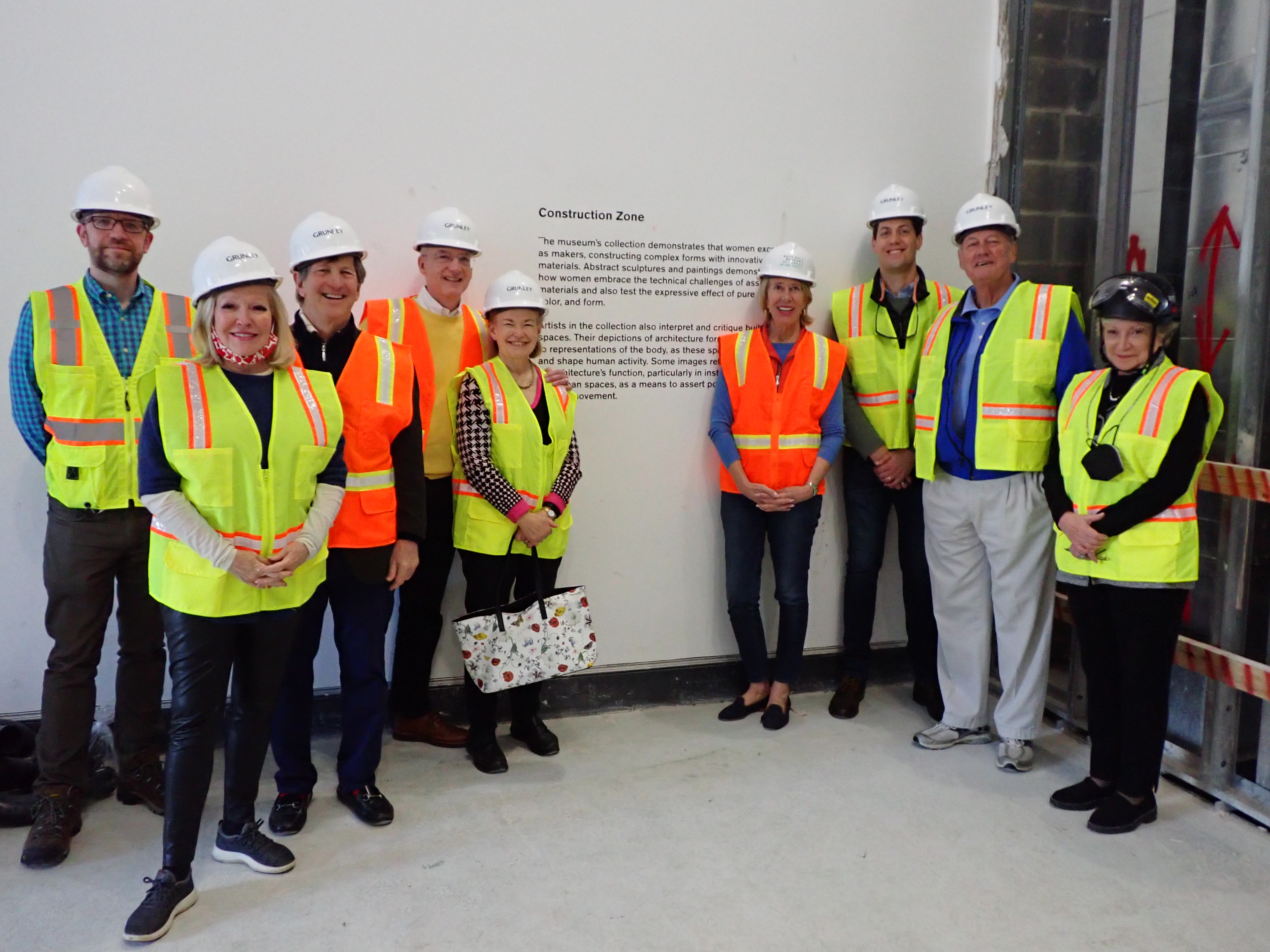 A group of 9 people wearing reflective vests and white hard hats standing in front of a white wall with text detailing about entering a construction zone.