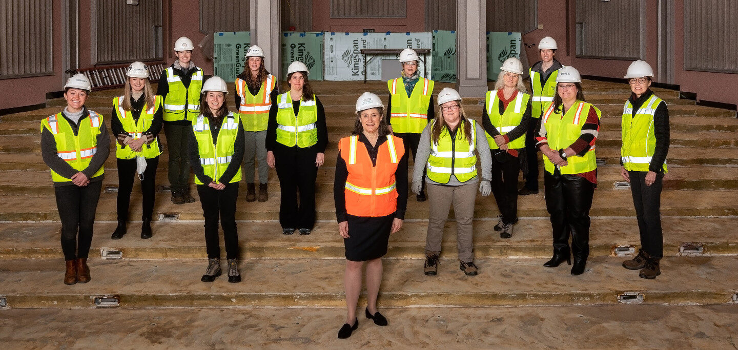 A group of 13 women wearing reflective vests and white hard hats standing in a large room under construction.