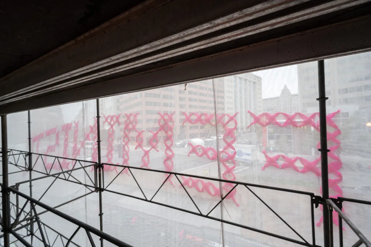 Interior view of construction scaffolding. The word "Feminist" is in large pink cross stitched letters on the outside of the scaffolding, seen backwards from this vantage point.