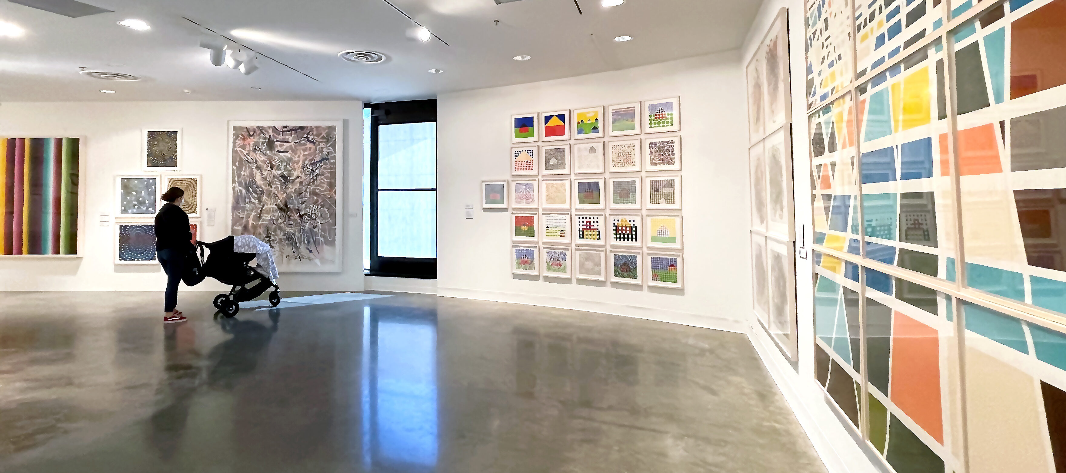 Woman pushes a stroller while looking at a variety of artworks of various sizes and colors in an exhibition gallery.