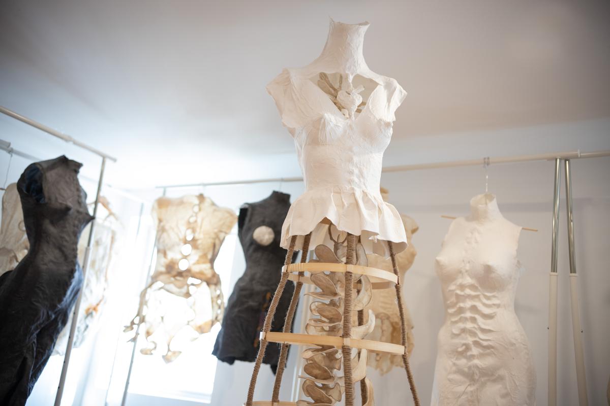 In a white-walled room six sculptural torsos are arranged, some hanging from an elevated rod, others standing on their own base. Two are made from black material. The others are white and differ slightly. Two seem to wear corsets, another shows spine-like ripples across the stomach. All have prominent breasts/nipples.