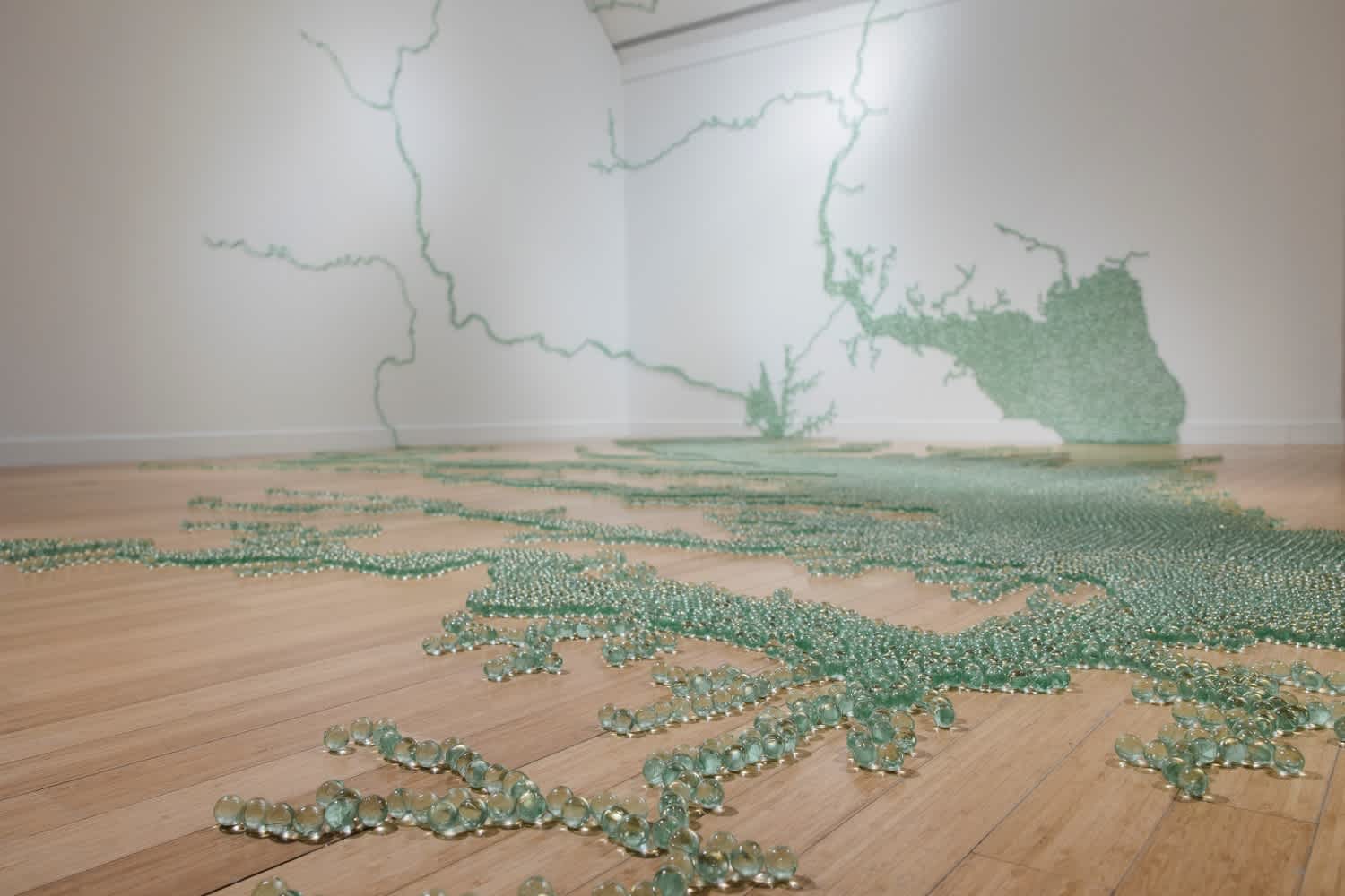 On the white walls and light wood floor of a gallery, thousands of small, sea-green glass marbles are affixed in patterns reminiscent of natural waterways as viewed from above.