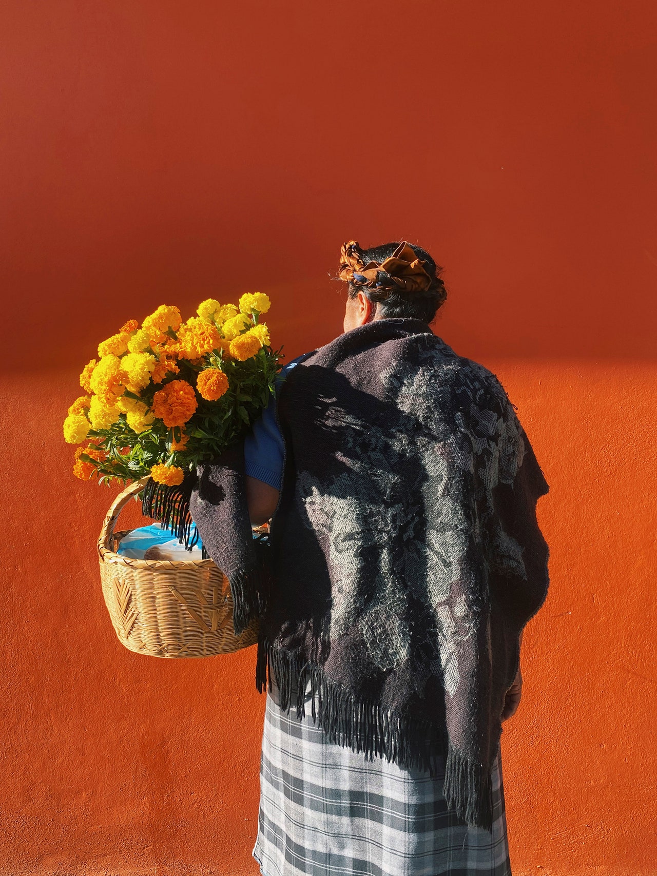 A color photograph features an Indigenous woman standing with her back to the camera in front of a bright orange wall. Its top half is in a shadow, and the bottom is in the sun. The woman holds a large bouquet of lush, yellow flowers and a light-tan woven basket. She wears a long, plaid skirt and a patterned shawl in grey. She looks slightly to the left, so that we see only her cheek and ear.