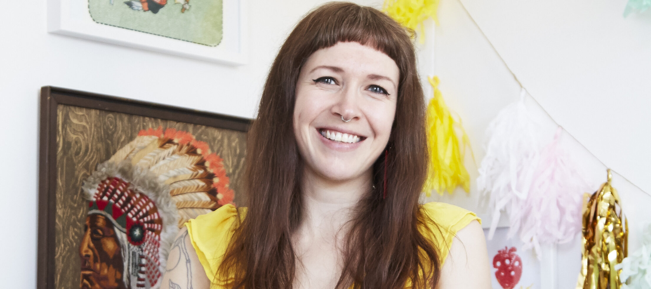 A light-skinned woman leans against a desk, smiling happily at the camera. She has straight, light-brown hair worn long to her breasts, with straight-cut bangs worn high on her forehead. She wears a yellow dress with ruffled sleeves and both arms have various tattoos. Behind her are various prints and artworks, most prominently an illustration of a Native American Chief in a headdress, shown in profile.