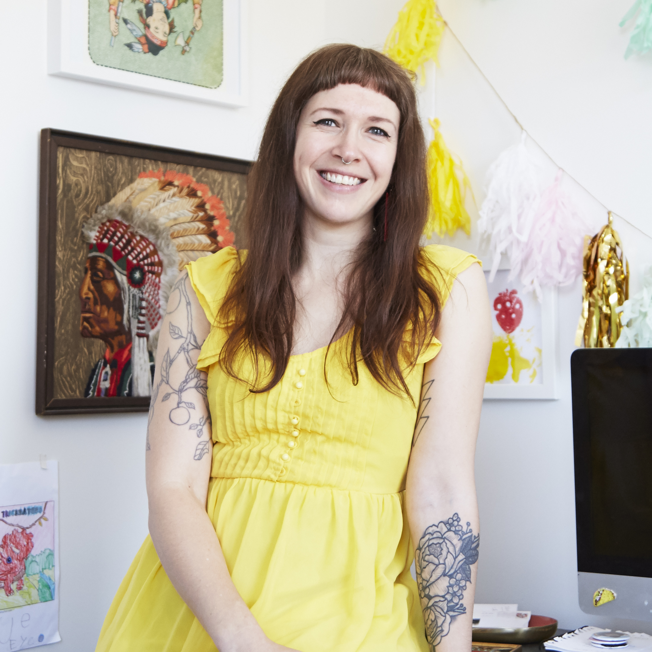 A light-skinned woman leans against a desk, smiling happily at the camera. She has straight, light-brown hair worn long to her breasts, with straight-cut bangs worn high on her forehead. She wears a yellow dress with ruffled sleeves and both arms have various tattoos. Behind her are various prints and artworks, most prominently an illustration of a Native American Chief in a headdress, shown in profile.