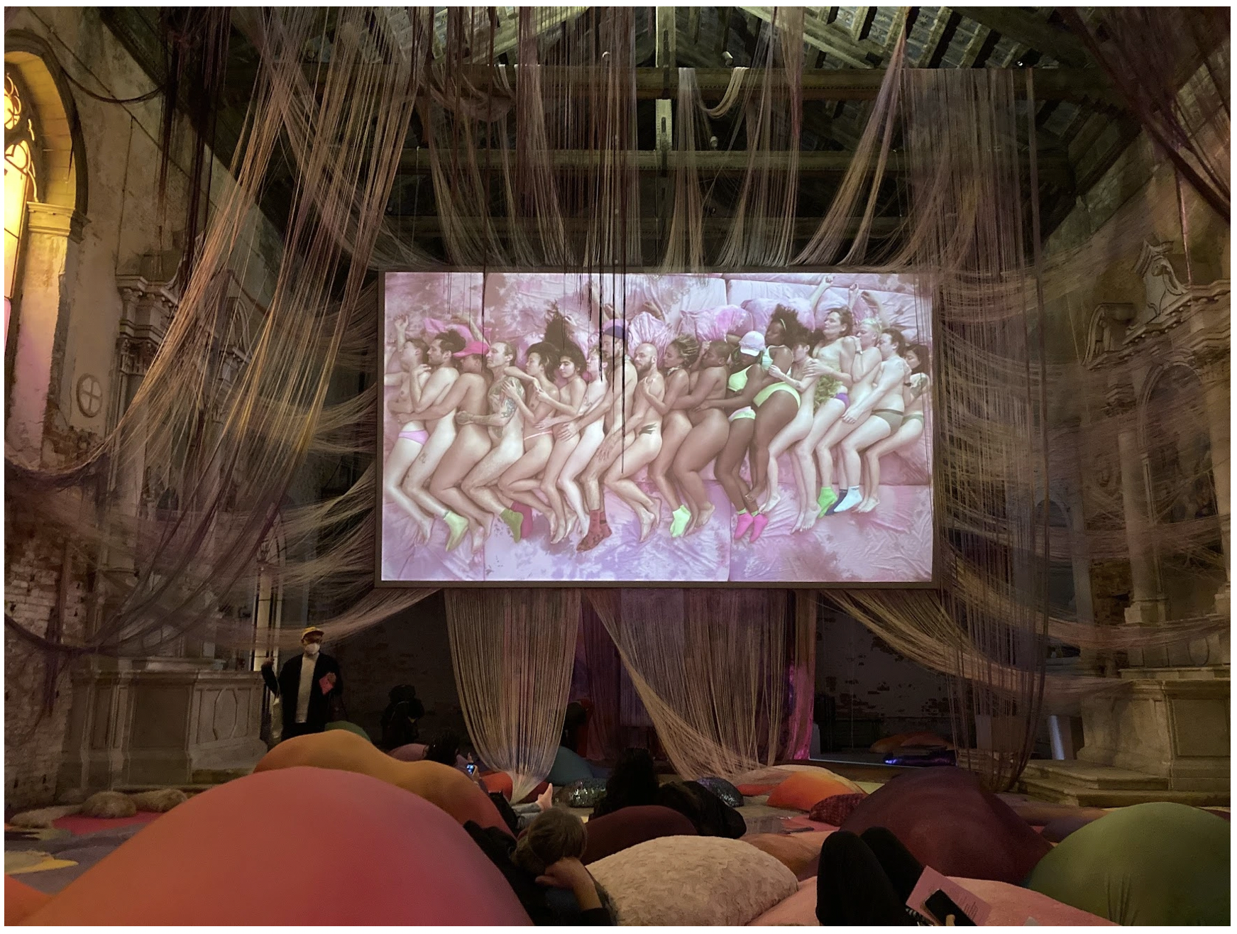 A large industrial hall is transformed into an art installation. From the high, cathedral ceilings, many strands of string or wire drapes down in various clusters around a large movie screen showing an image of a line of naked bodies all spooning one another stop pink sheets. Participants are diverse in ethnicity and gender. Below this screen, viewers recline on soft, bulbous, multi-colored pillows that are arranged on the floor.