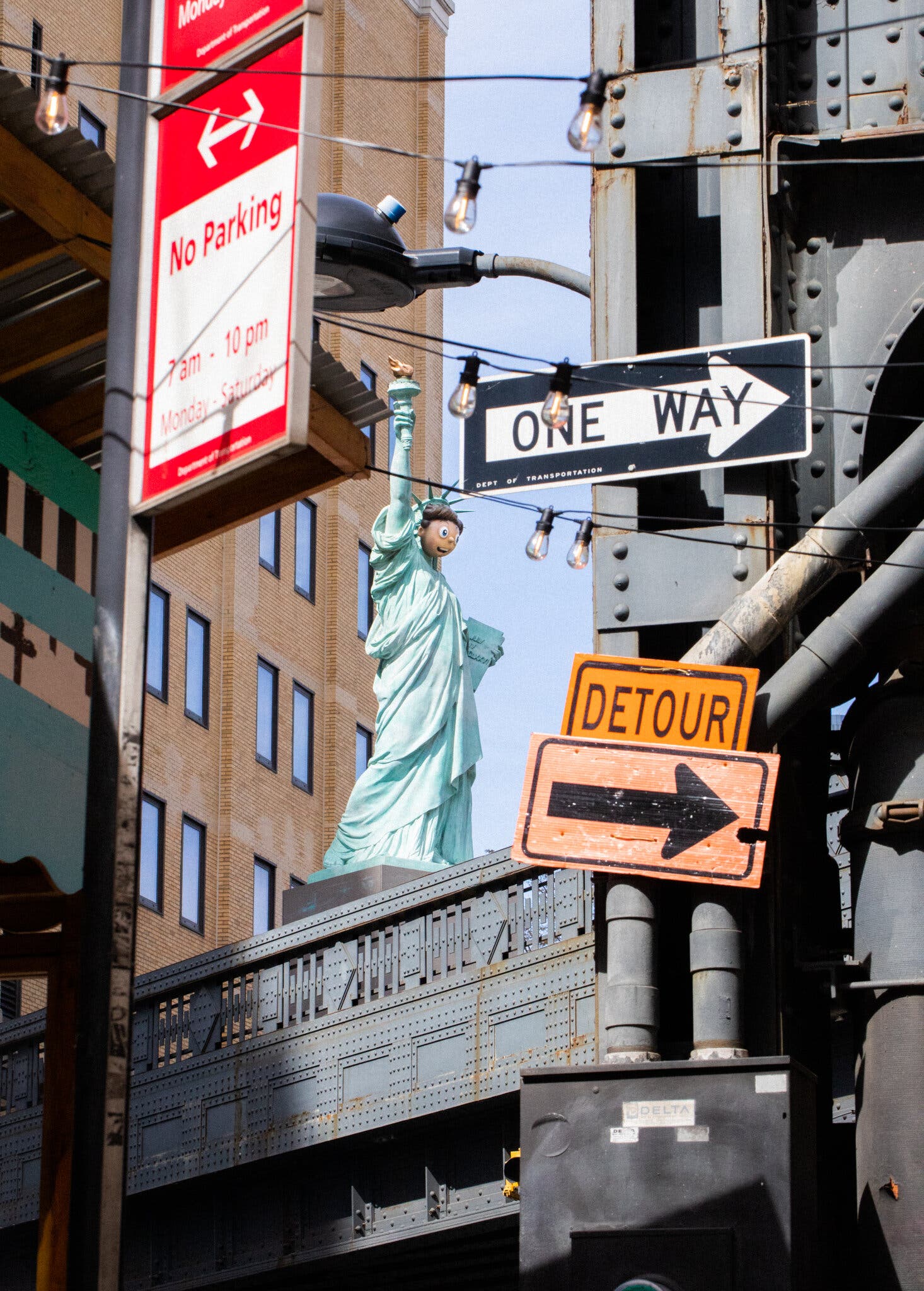 Visible at a distance through a busy city landscape of traffic and parking signs, a tall building, and a lamppost, a large sculputral figure of the Statue of Liberty stands on a pedestal on top of a walkway or bridge. It's face is that of a boy-like cartoon emoji.