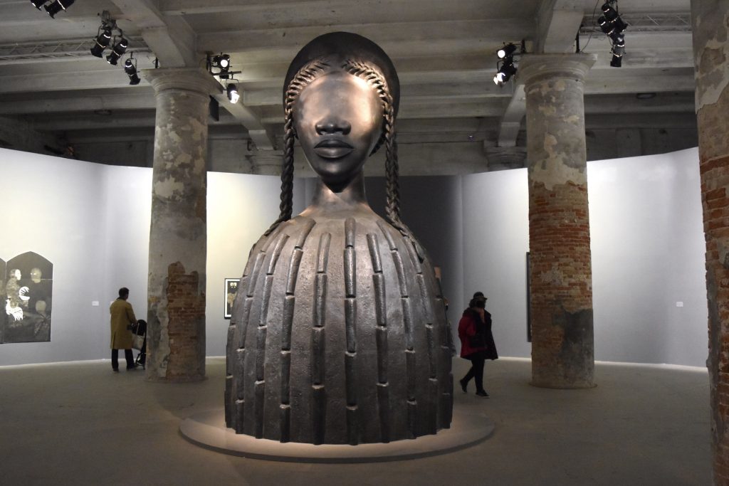 A 16-foot-high bronze sculpture sits in the middle of an industrial gallery space. It features the head of a woman, sans eyes, atop a cylandrical shape. She has two long braids on either side of her head. The sculpture is sphinx-like.