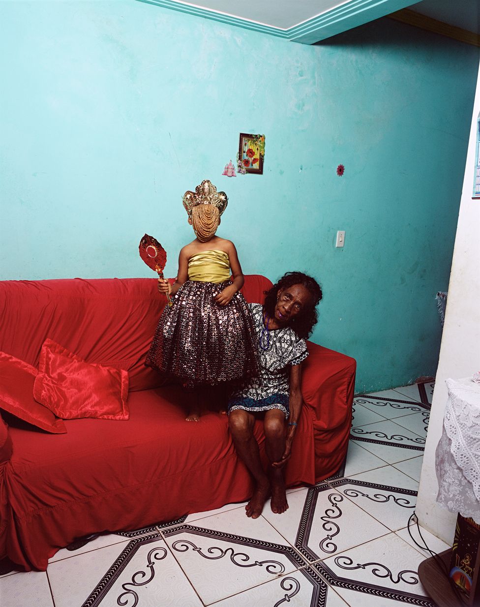 An older, dark-skinned woman sits on the end of a couch draped in red fabric. Her left hand reaches down to cup her calf, her right is positioned behind a young girl, who stands on the cushion next to her wearing a sparkly costume skirt, gold tube top, and a crown with beaded detail that obscures her face. The girl also holds a regal, antique mirror in her left hand. The room they are in has sparse, bright blue walls that are marked with stains and wear and a tiled floor.