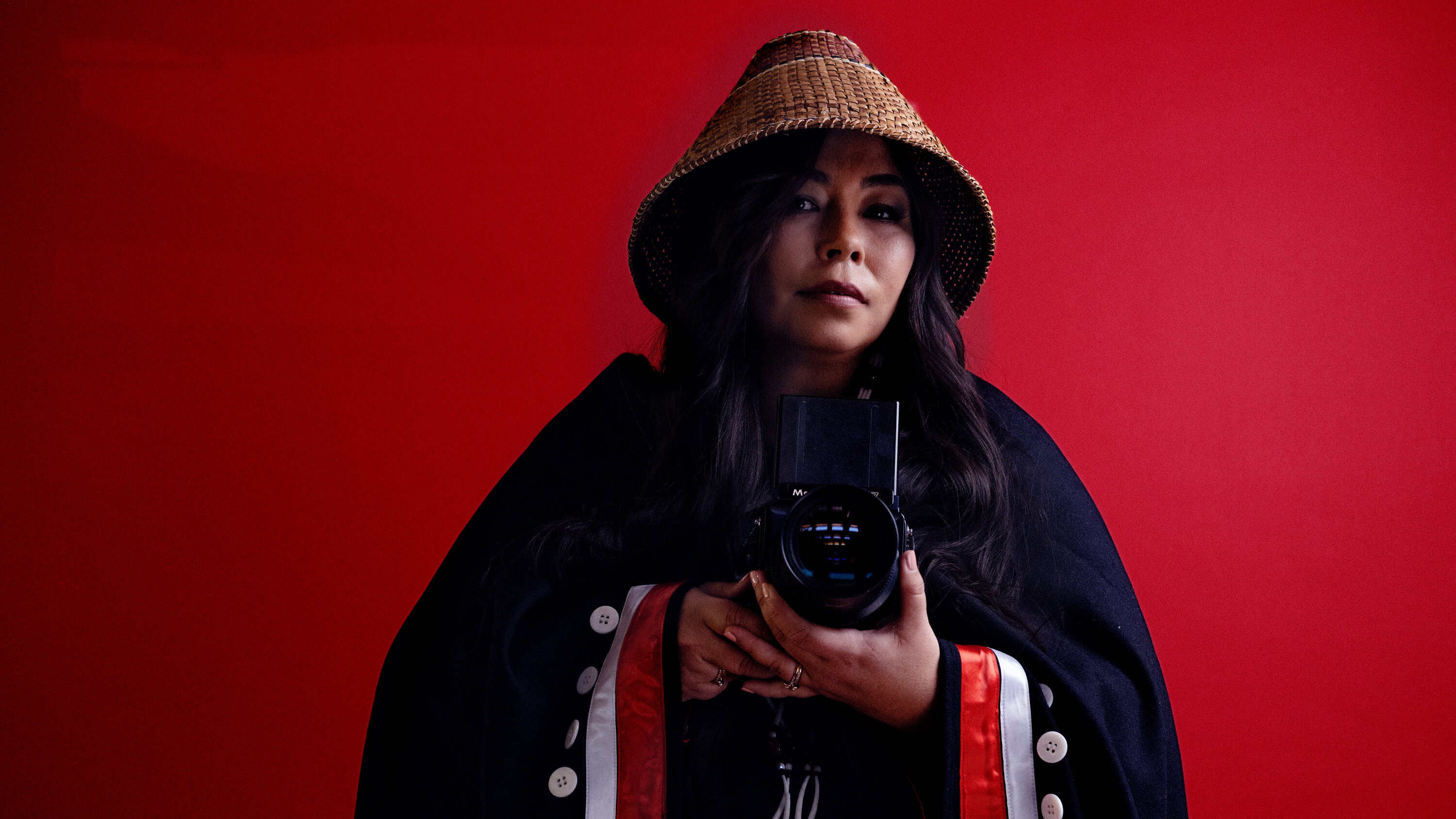 Photographer Matika Wilbur takes a mirror selfie with a large, black SLR camera before a bright red background. She wears a straw hat, a straw belt, and a black coat draped over her like a cape embroidered with white buttons and white and red ribbons.