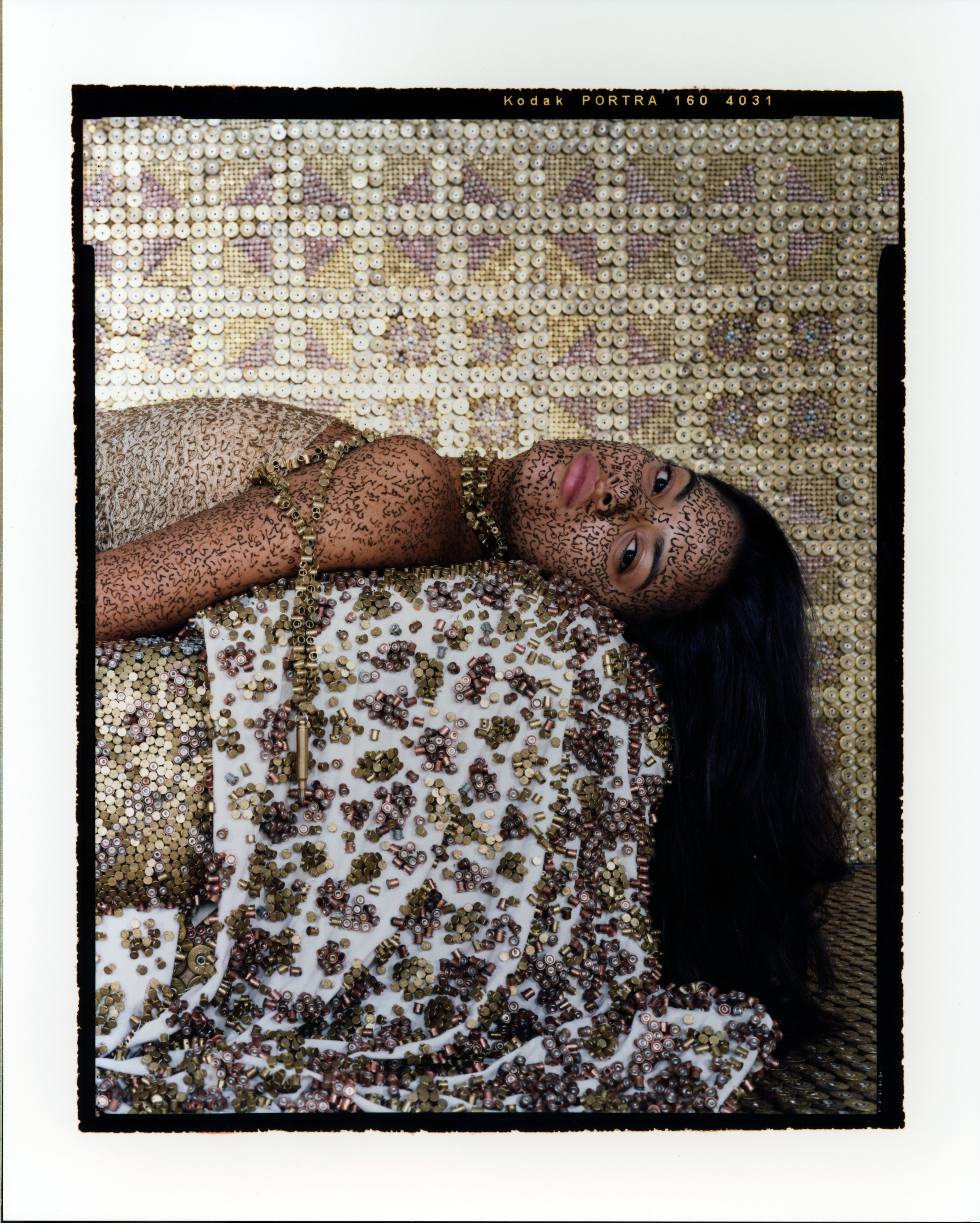 A woman lying on her back on a bed, seen from the chest up. Her head turns left to look at the viewer. She wears a light, sleeveless top with long necklaces and her long, dark hair falls to the floor. A metallic surface covers everything—the walls, furniture, and the woman.