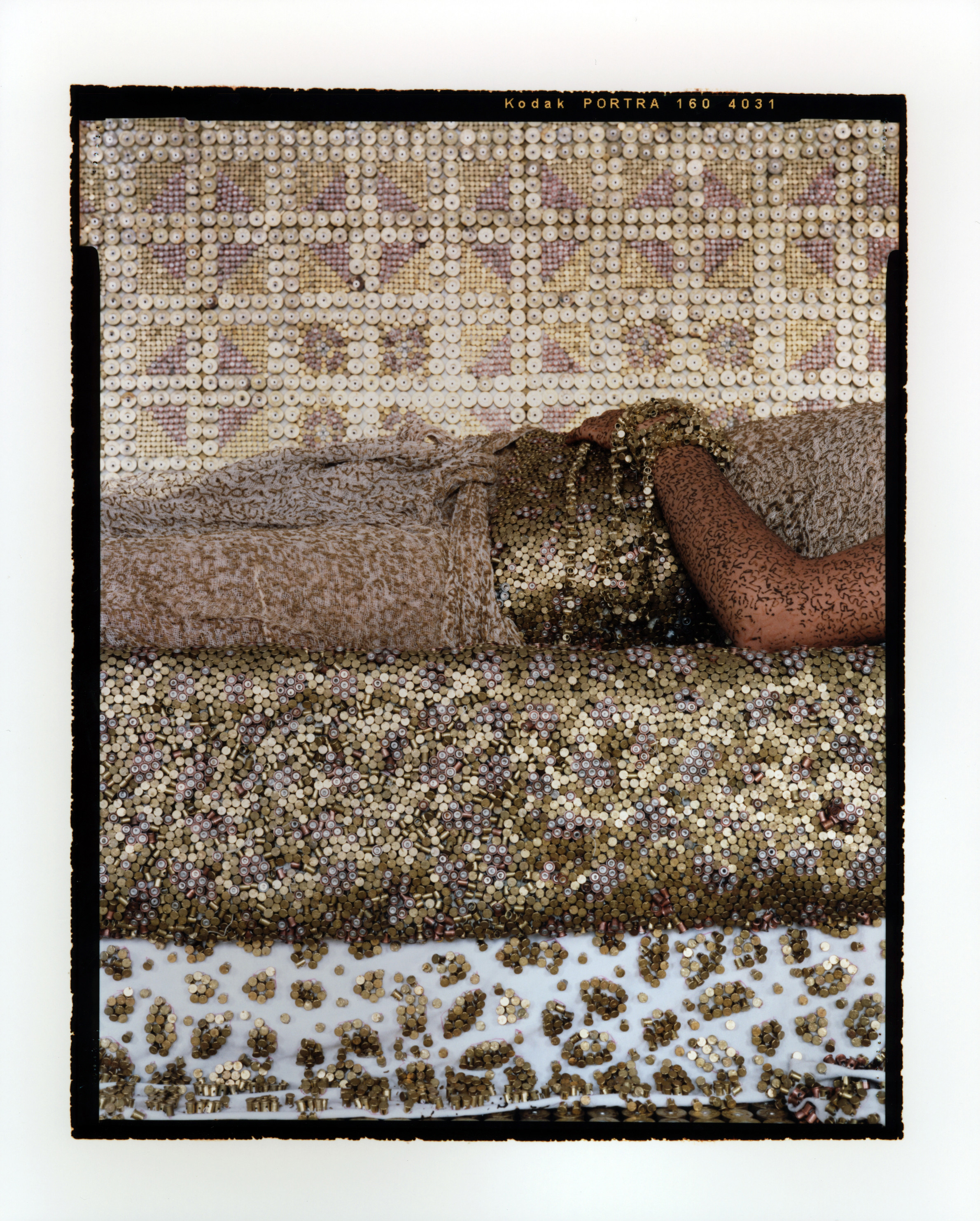 The mid-section of a medium-light skinned adult woman lying on her back on a bed, seen from the side. She wears a light, airy dress and a wide, golden belt and bracelets. A metallic surface covers everything—the walls, furniture, and the woman.