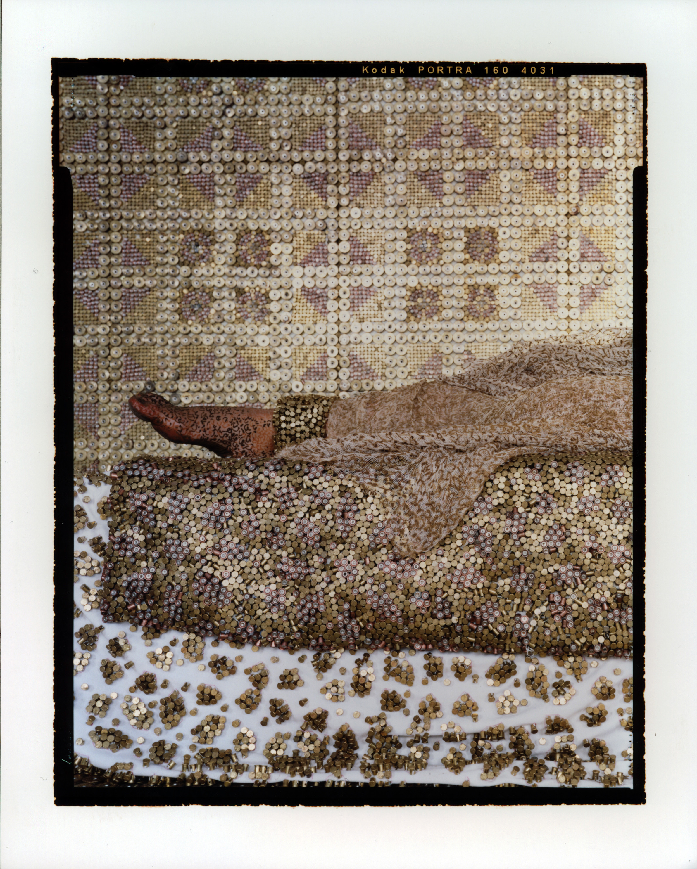 The  legs of a medium-light skinned adult woman lying on her back on a bed, seen from the side from the knees down. A light, airy dress covers her legs to the ankles, leaving her feet bare. A metallic surface covers everything—the walls, furniture, and the woman.