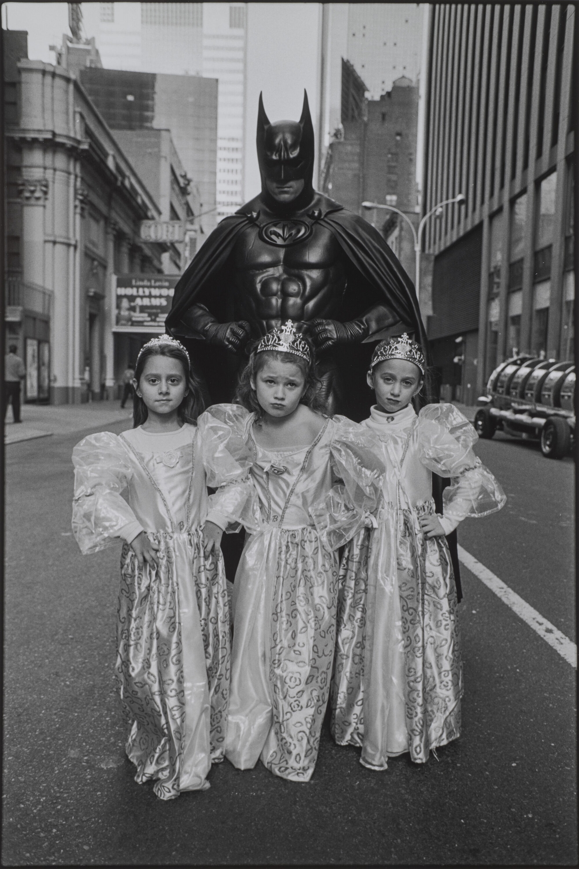 A black-and-white photograph of three light-skinned young girls in princess costumes of dresses and crowns standing in front of an adult in a full Batman costume. They all stand in the middle of a city street with defiant poses, their hands on their hips and faces serious.
