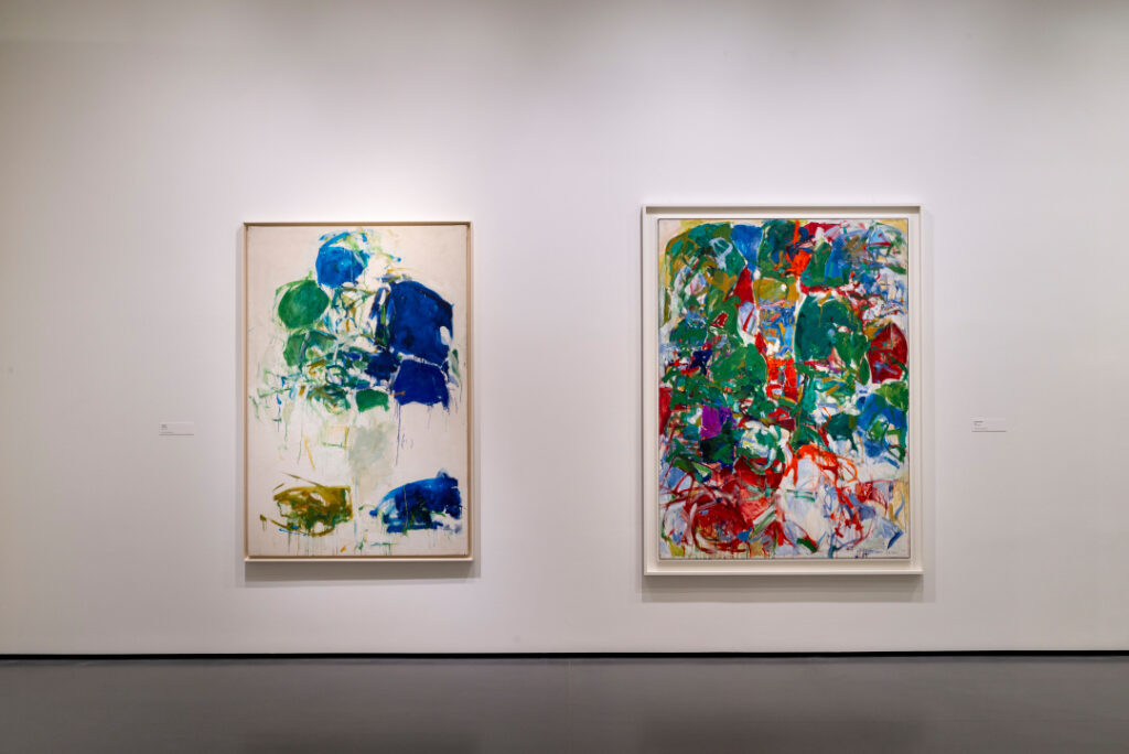 An exhibition view shows two abstract expressionist paintings hanging on a white wall. The brushstrokes are energic, dynamic, and erratic. On the painting on the left, the prominent colors are blue, green, and white. The painting on the right is more chaotic, including a range of colors: green, red, yellow, blue, white, and purple.