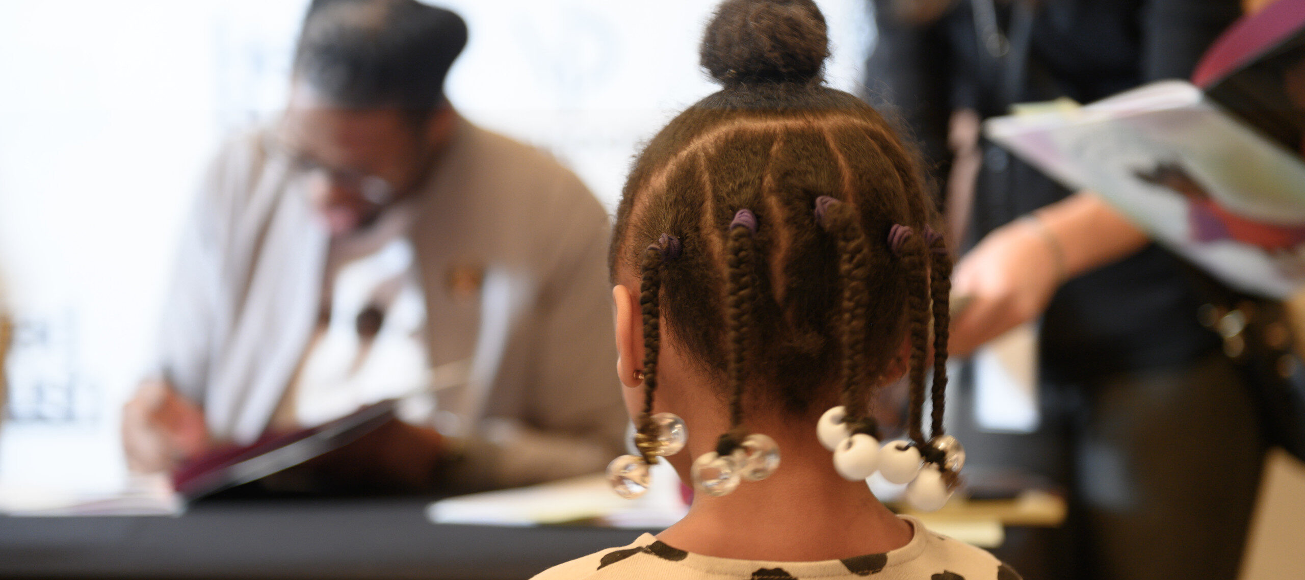 Back of a young girl's head with five braids and a bun on top. She wears a white shirt with black polka dots and is standing waiting for a book signing.