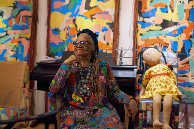 An elderly African American woman with long, grey braids is photographed amidst her artworks in her studio, smiling. Sitting on a chair before three large-scale paintings and next to a life-size textile doll, she wears a colorful, quilted dress and a deep-blue headband.