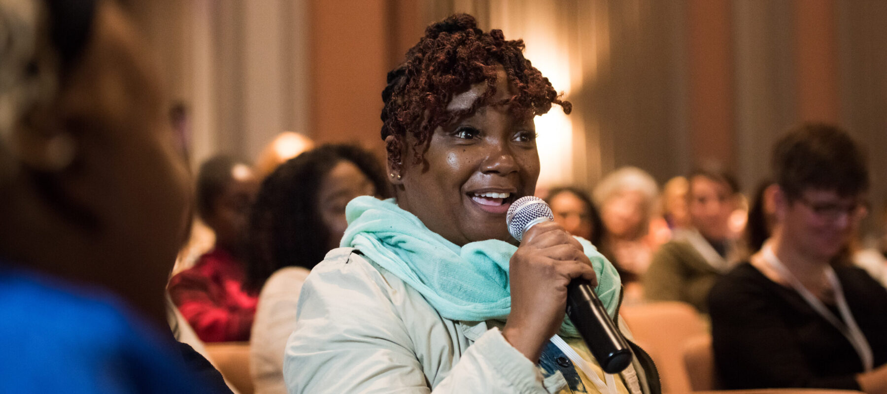 A dark-skinned woman with short auburn dreadlocks sits in an auditorium. She is speaking into a microphone with an enthusiastic expression on her face.