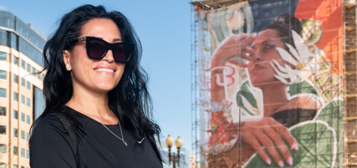 A woman with medium skin tone with long dark hard, sunglasses, and a black shirt stands in front of the exterior of the museum and the large, colorful mural on the front.