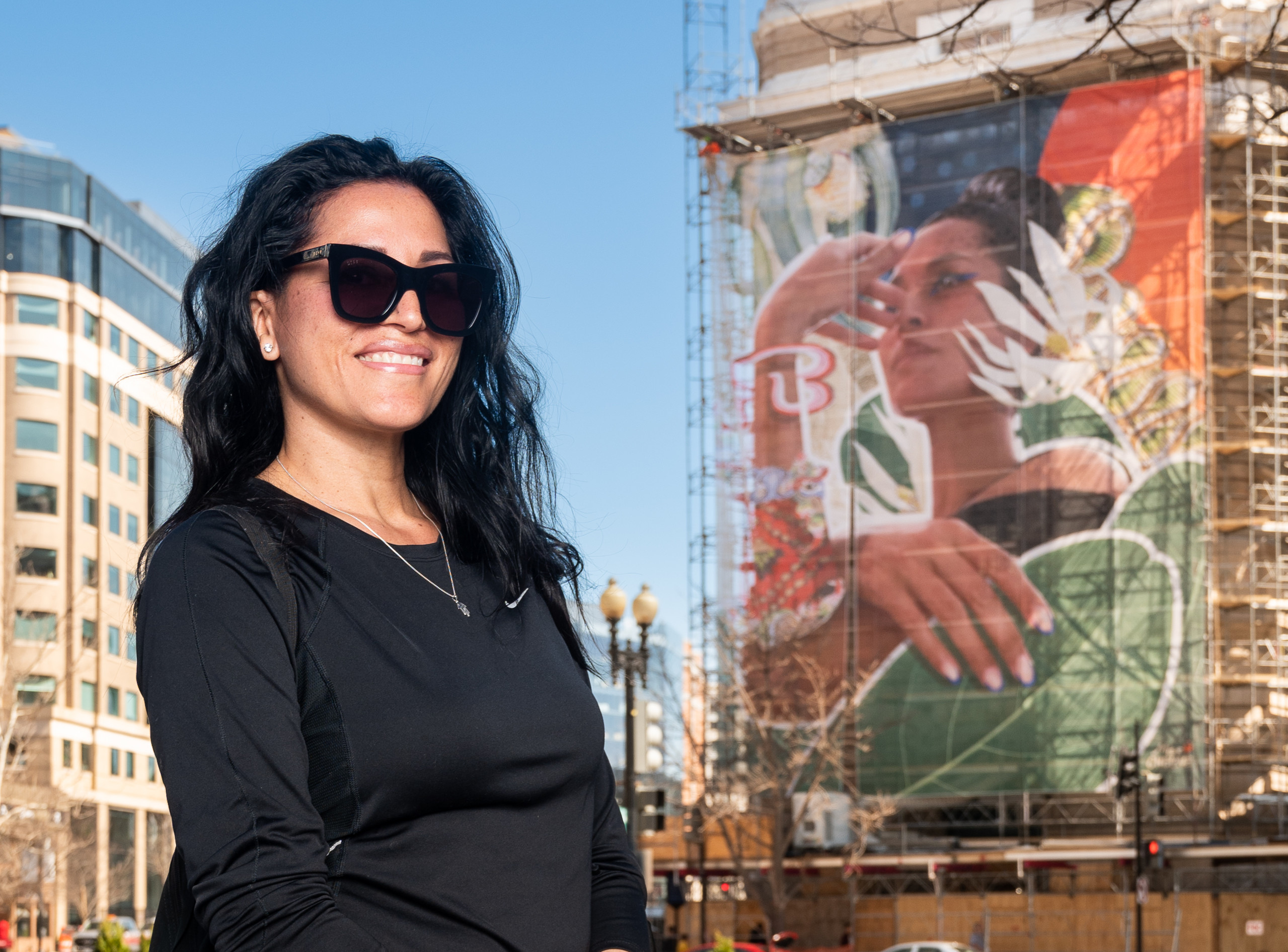 A woman with medium skin tone with long dark hard, sunglasses, and a black shirt stands in front of the exterior of the museum and the large, colorful mural on the front.