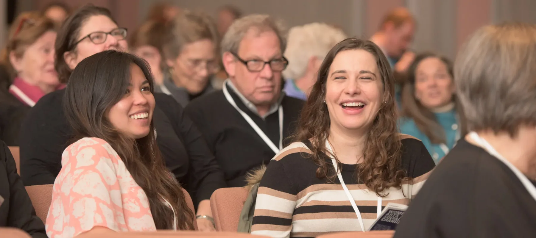 Two light-skinned women with long brown hair sit in a full auditorium. They are both laughing.