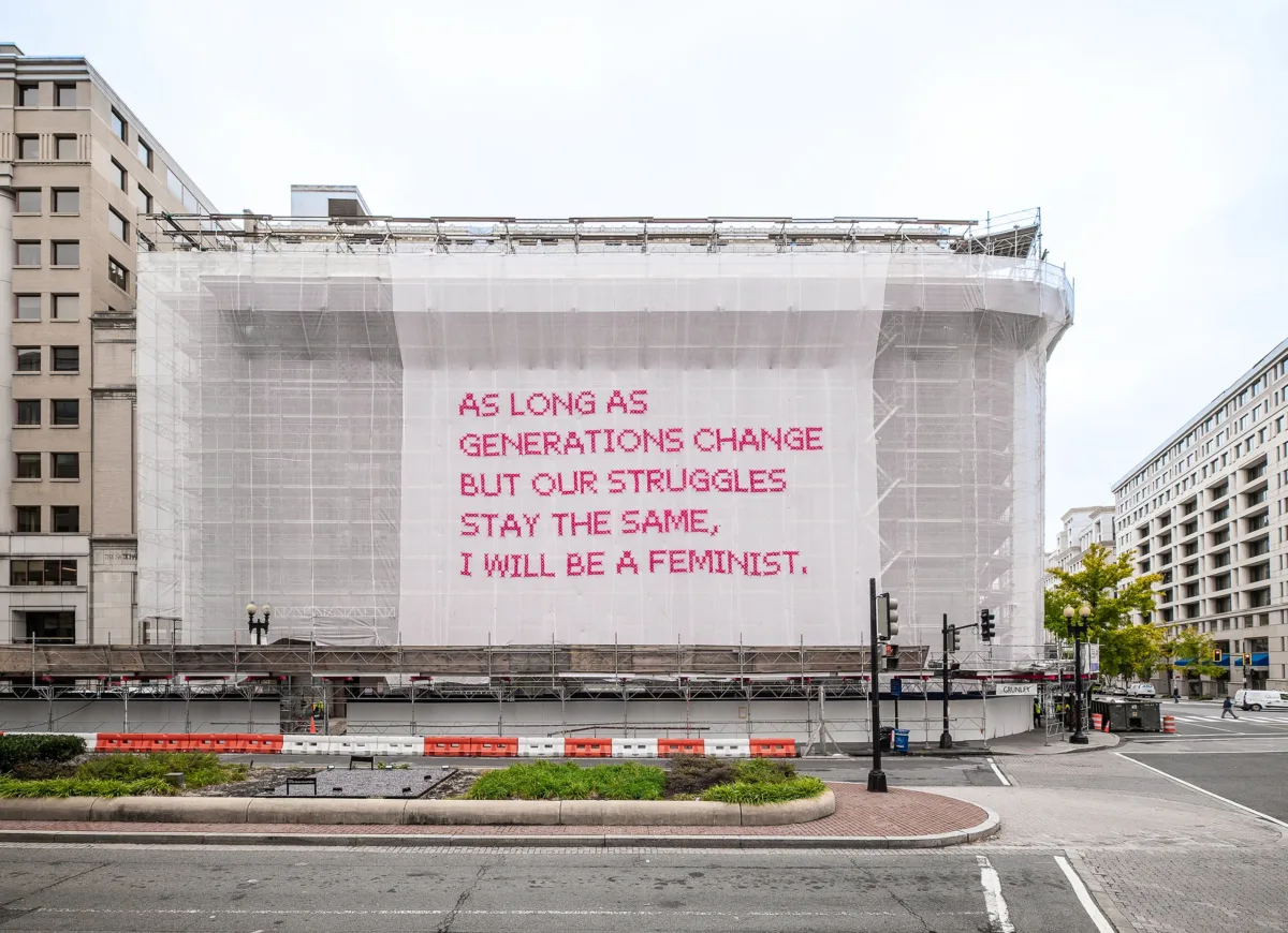 White gauzy fabric hangs in front of scaffolding surrounding a city building. Bold magenta letters on the fabric read, “As long as generations change but our struggles stay the same, I will be a feminist.”