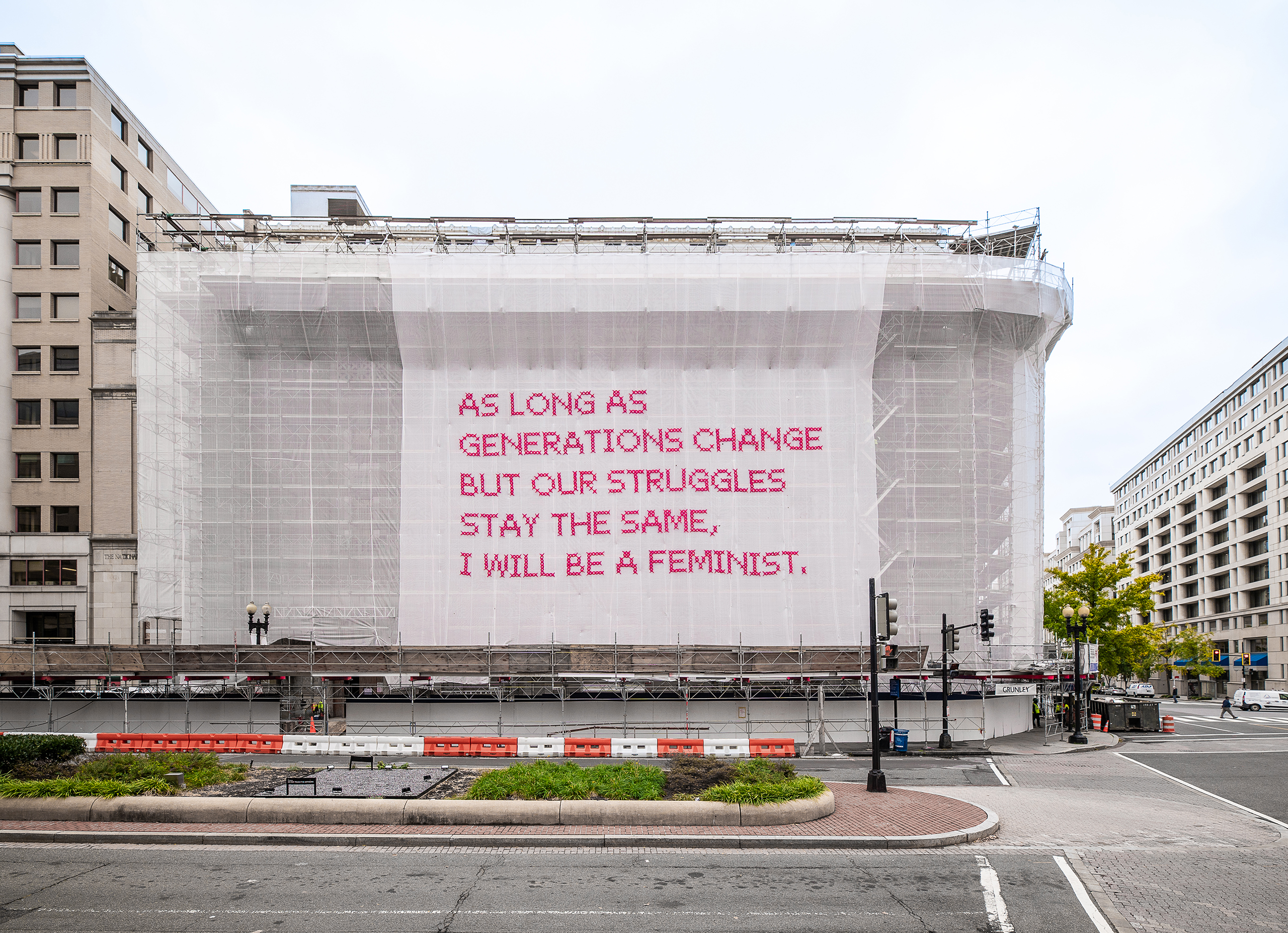 White gauzy fabric hangs in front of scaffolding surrounding a city building. Bold magenta letters on the fabric read, “As long as generations change but our struggles stay the same, I will be a feminist.”