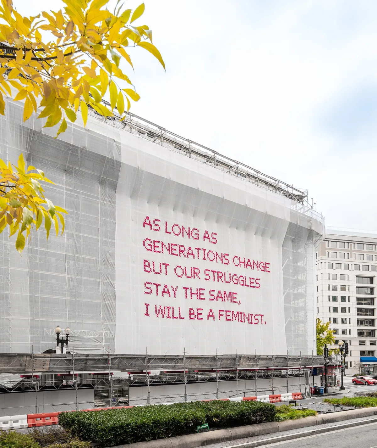 White gauzy fabric hangs in front of scaffolding surrounding a city building. Bold magenta letters on the fabric read, “As long as generations change but our struggles stay the same, I will be a feminist.”