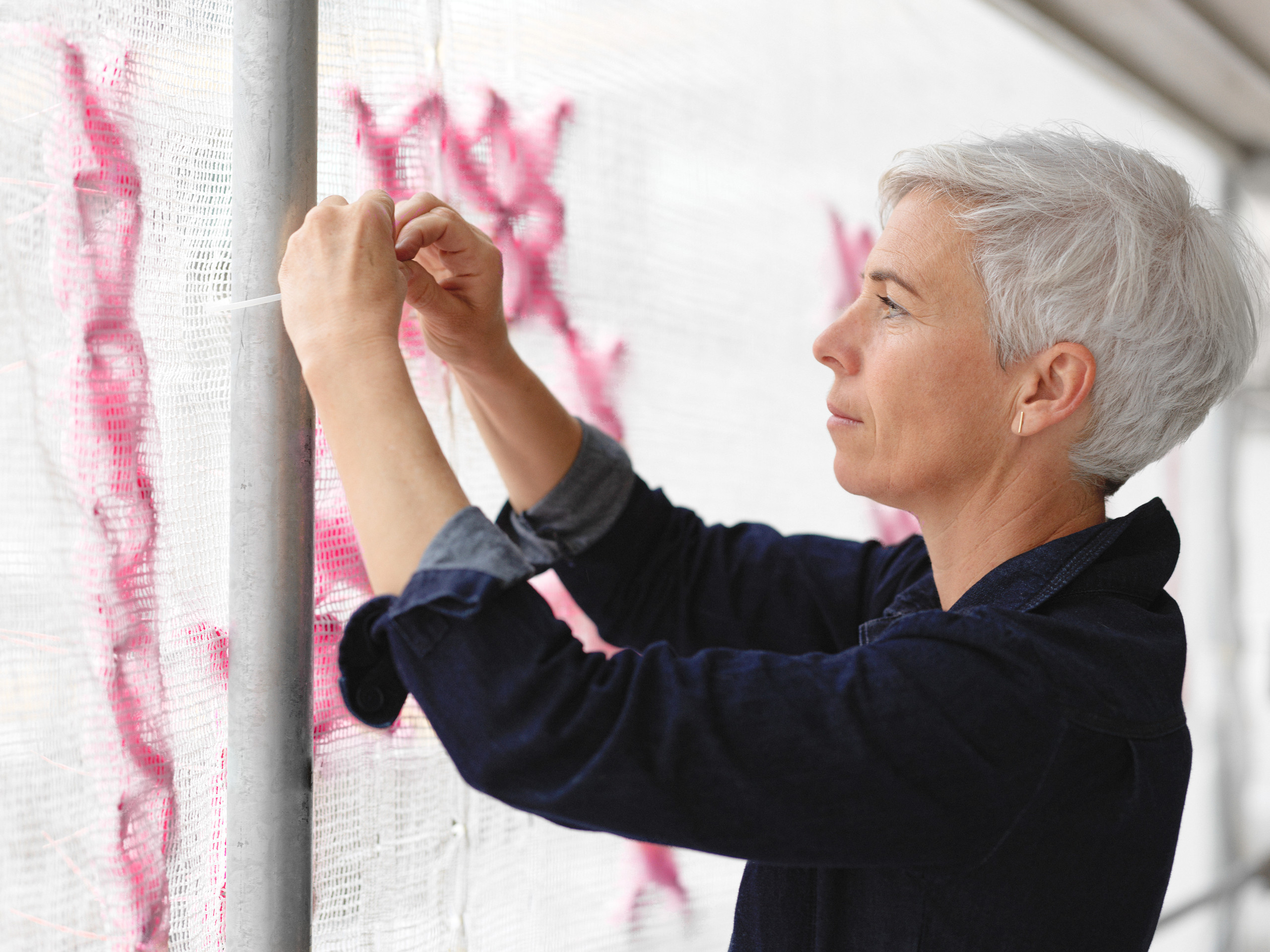 A light-skinned white woman with short white hair stands in profile, tying white scrim with bright pink stitching to a metal pole.