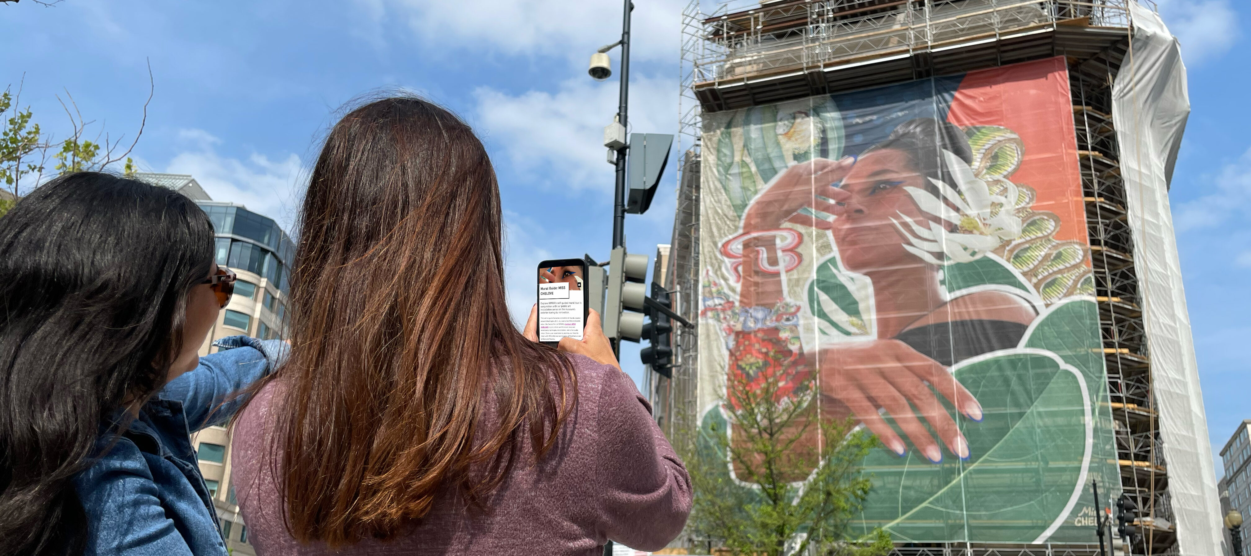 Two women with medium skin tone and long brown hair use their cell phone to look at the online mural guide while looking at the large mural on the museum's building exterior.