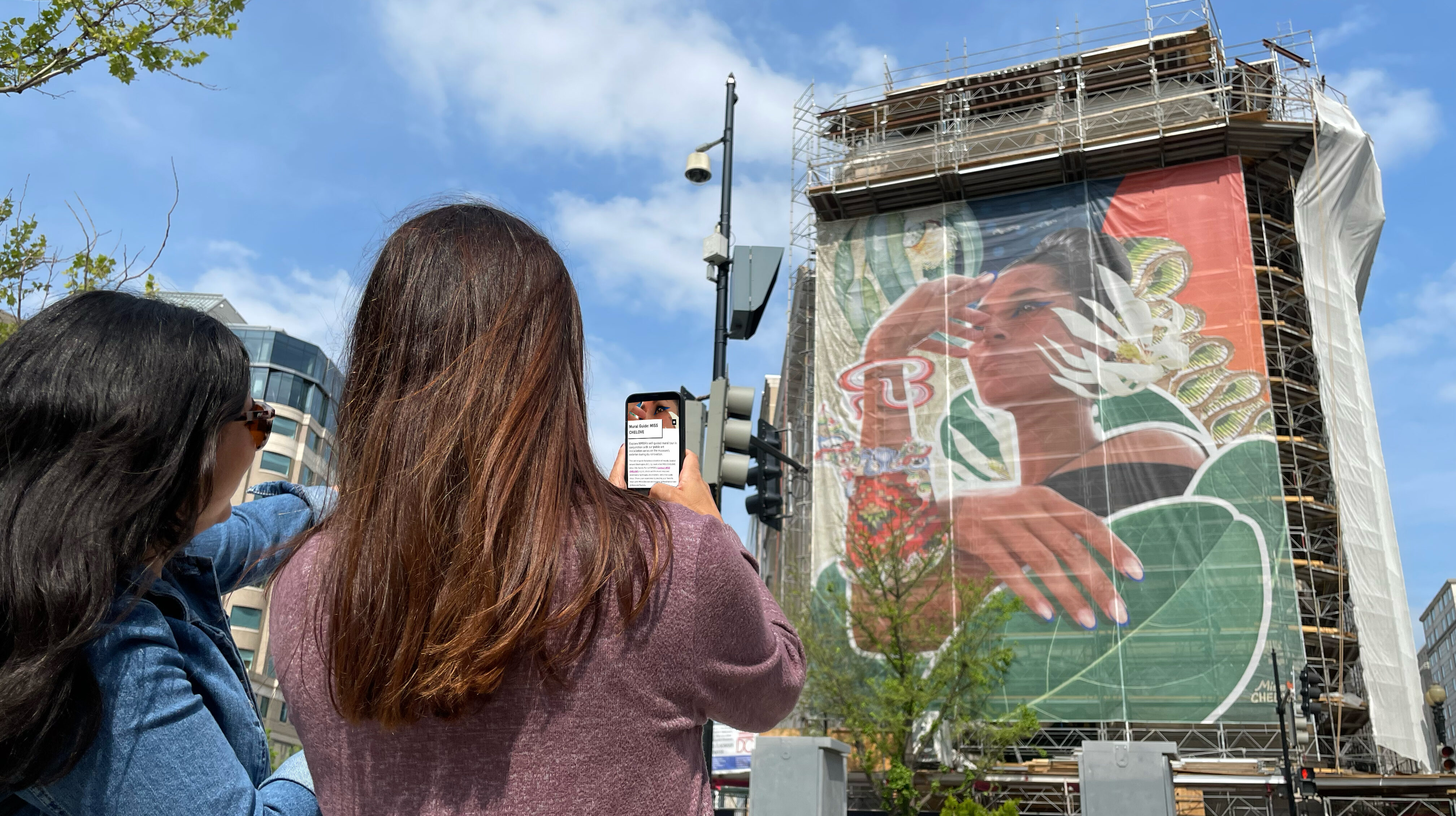 Two women with medium skin tone and long brown hair use their cell phone to look at the online mural guide while looking at the large mural on the museum's building exterior.