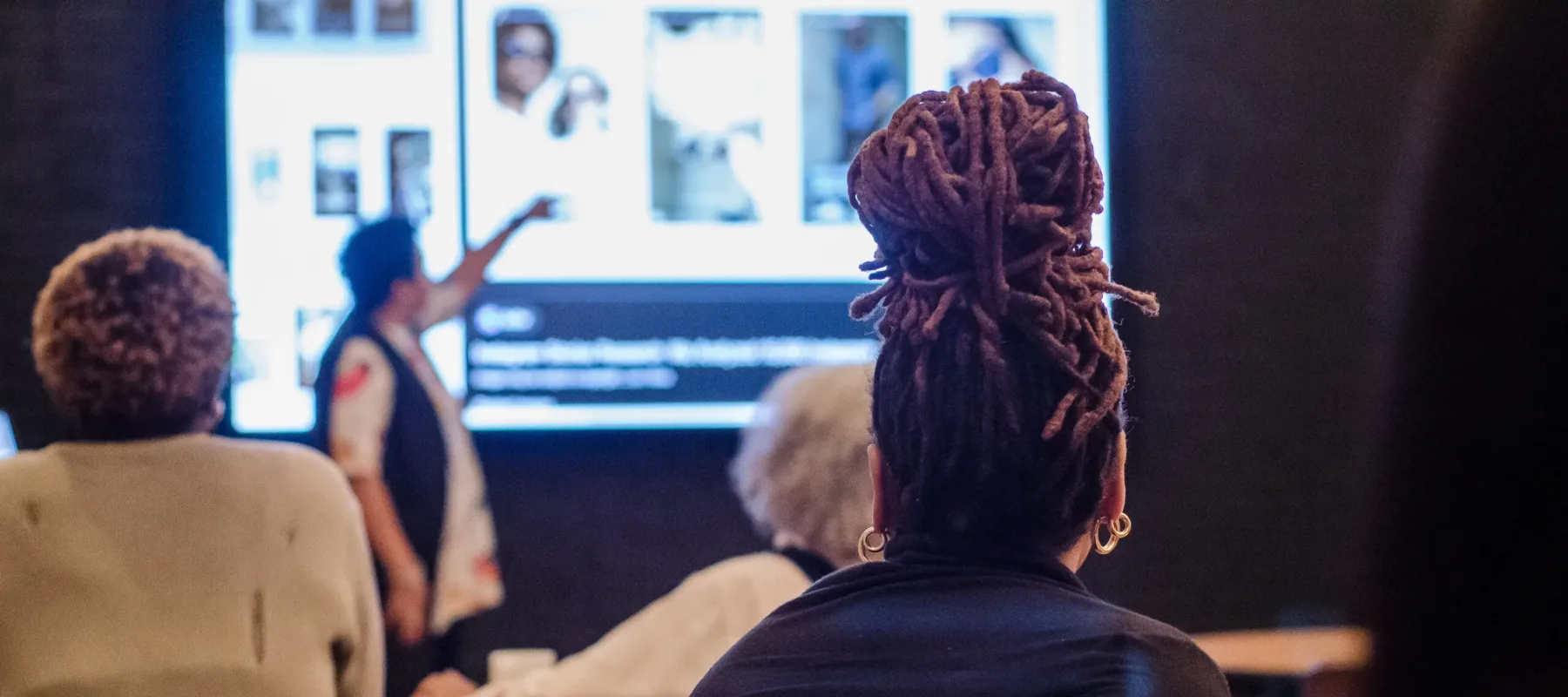 Three adults sitting and looking at a facilitator pointing at a presentation during a workshop.