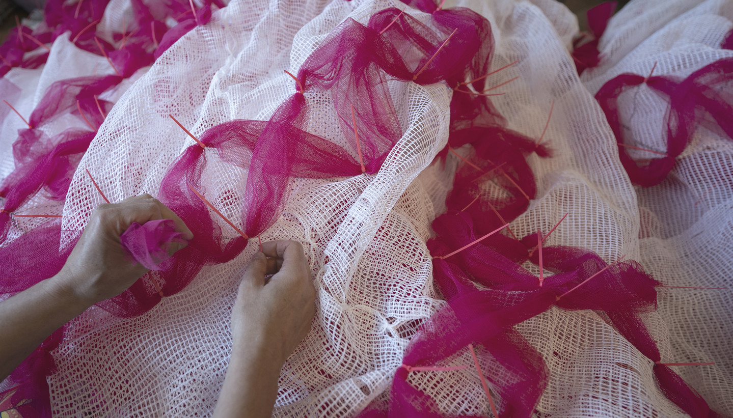 A close-up photograph of two light-skinned hands sewing bright pink tulle onto white mesh.