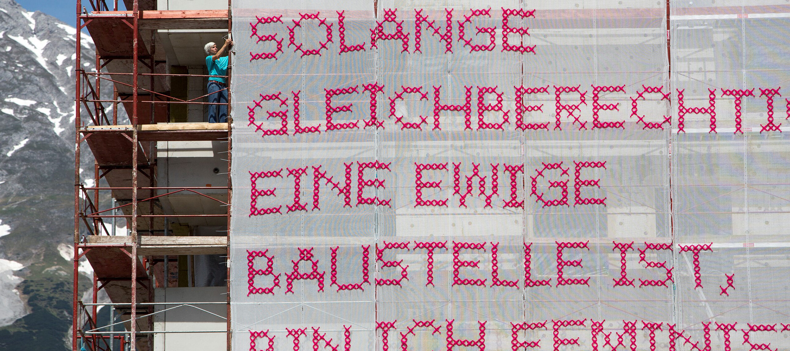 A light-skinned woman with short, platinum blonde hair stands on scaffolding attaching a very large panel of white gauzy material with “Solange Gleichberechtigung eine ewige Baustelle ist, bin ich Feministin” stitched in bright pink lettering to the bars of the scaffold. In the background you can see a bight blue sky and snow capped mountain.
