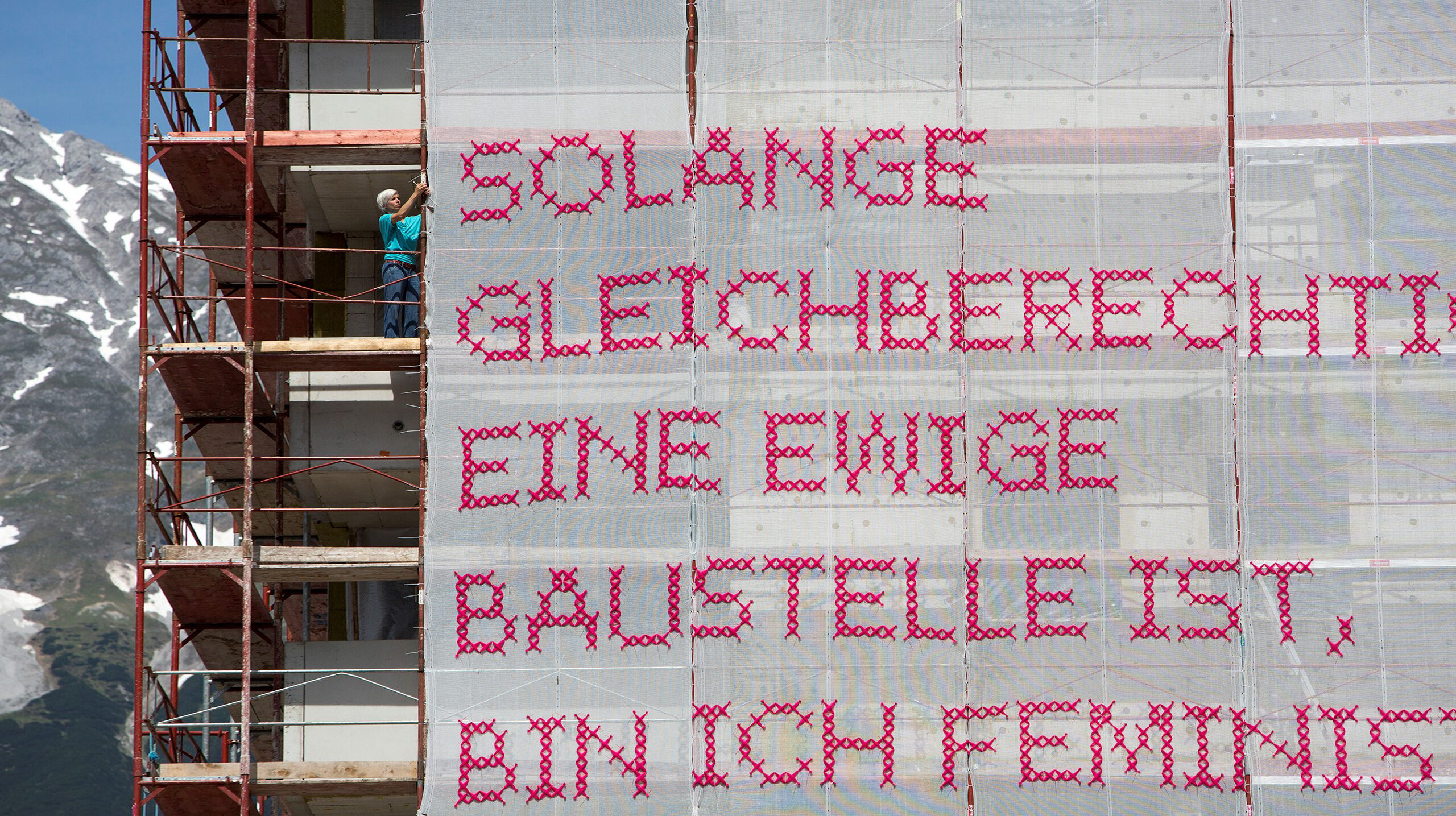 A light-skinned woman with short, platinum blonde hair stands on scaffolding attaching a very large panel of white gauzy material with “Solange Gleichberechtigung eine ewige Baustelle ist, bin ich Feministin” stitched in bright pink lettering to the bars of the scaffold. In the background you can see a bight blue sky and snow capped mountain.