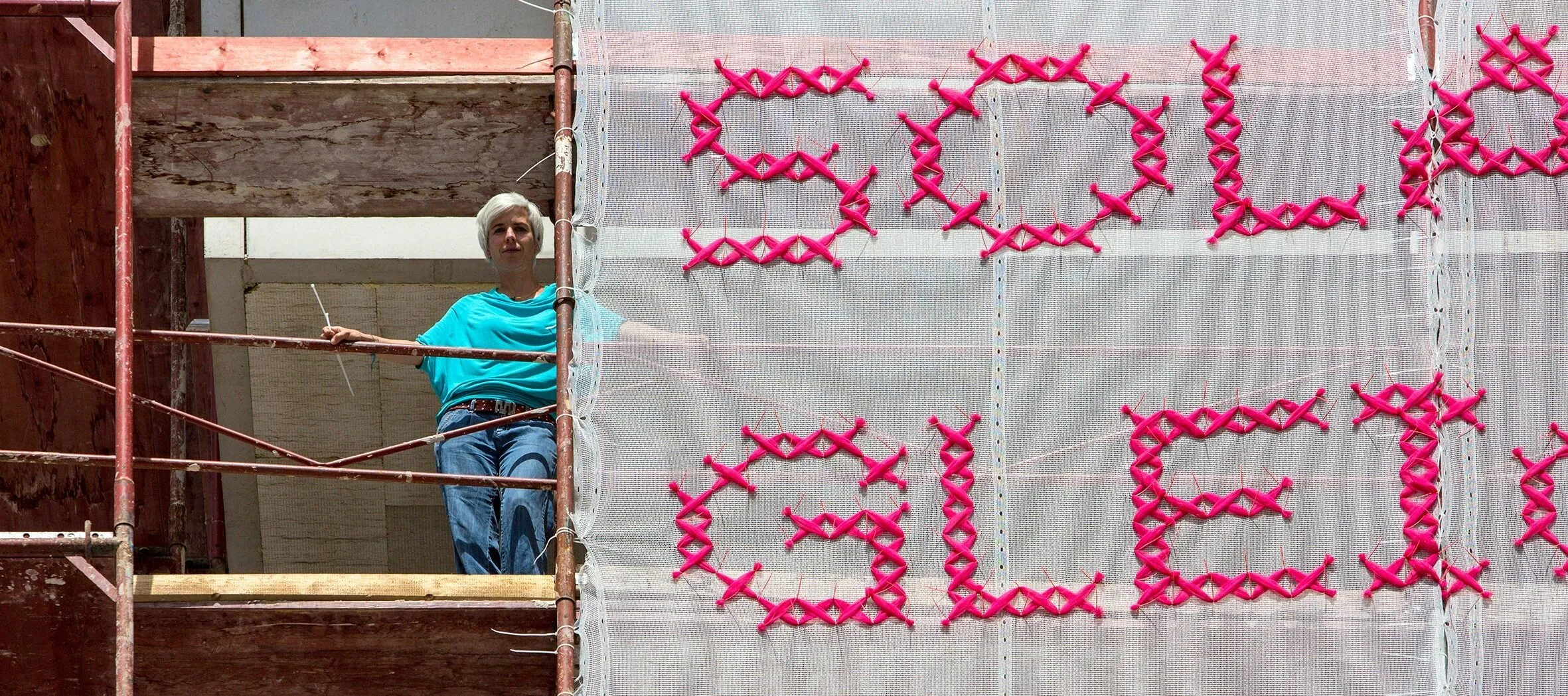 A light-skinned woman with short, platinum blonde hair stands on scaffolding next to a very large panel of white gauzy material with “Solange Gleichberechtigung eine ewige Baustelle ist, bin ich Feministin” stitched in bright pink lettering.