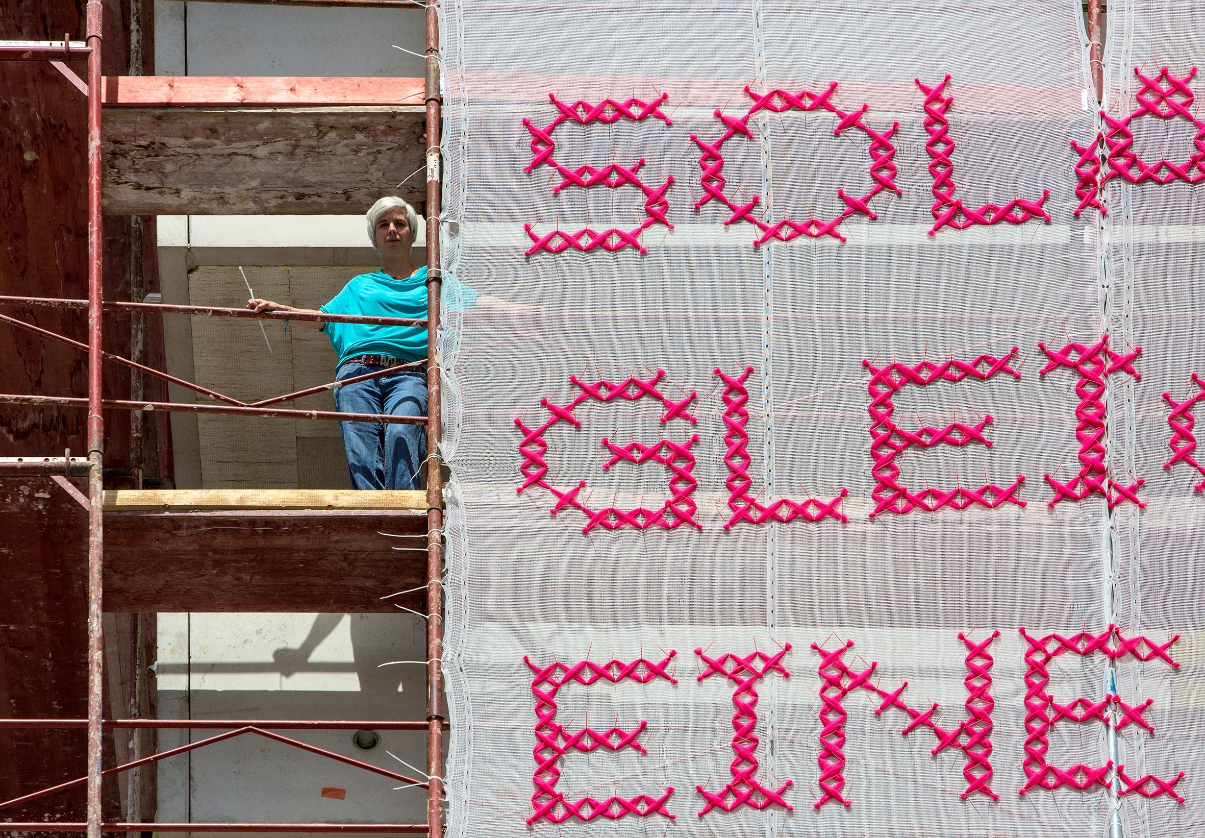 A light-skinned woman with short, platinum blonde hair stands on scaffolding next to a very large panel of white gauzy material with “Solange Gleichberechtigung eine ewige Baustelle ist, bin ich Feministin” stitched in bright pink lettering.