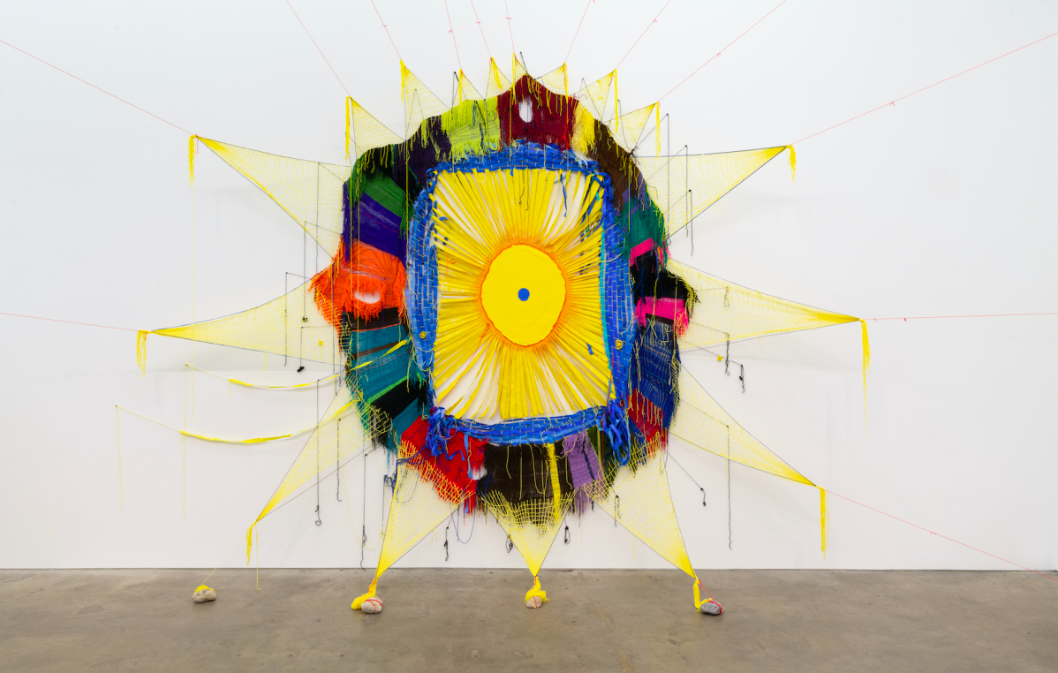 Against a white gallery wall, one of Michelle Sagre’s yarn installations is presented. A variety of materials and textures form a large, colorful, star-like shape. It is held up from all of its points by strings. 