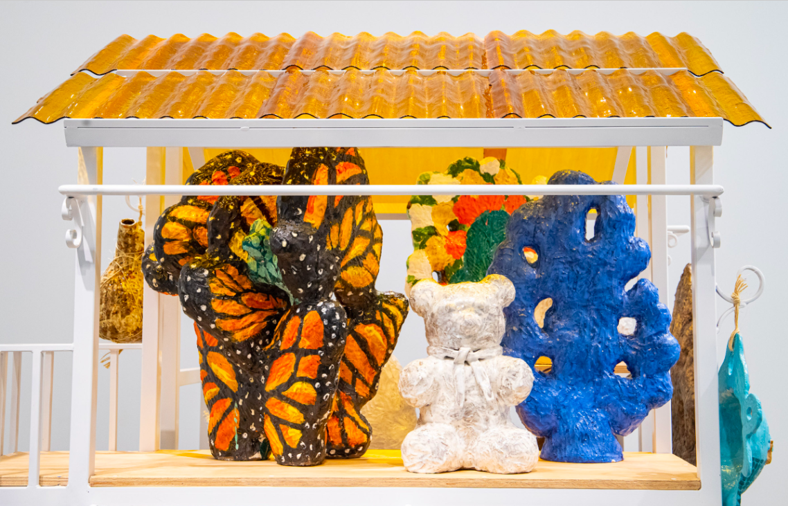 Three sculptures stand on a construction made out of metal, wood, and glass. The construction mimics the rooftop of a house with its yellow stained glass leaving a golden shimmer upon the sculptures. The sculptures have the form of a butterfly, a teddy bear, and a coral reef. 